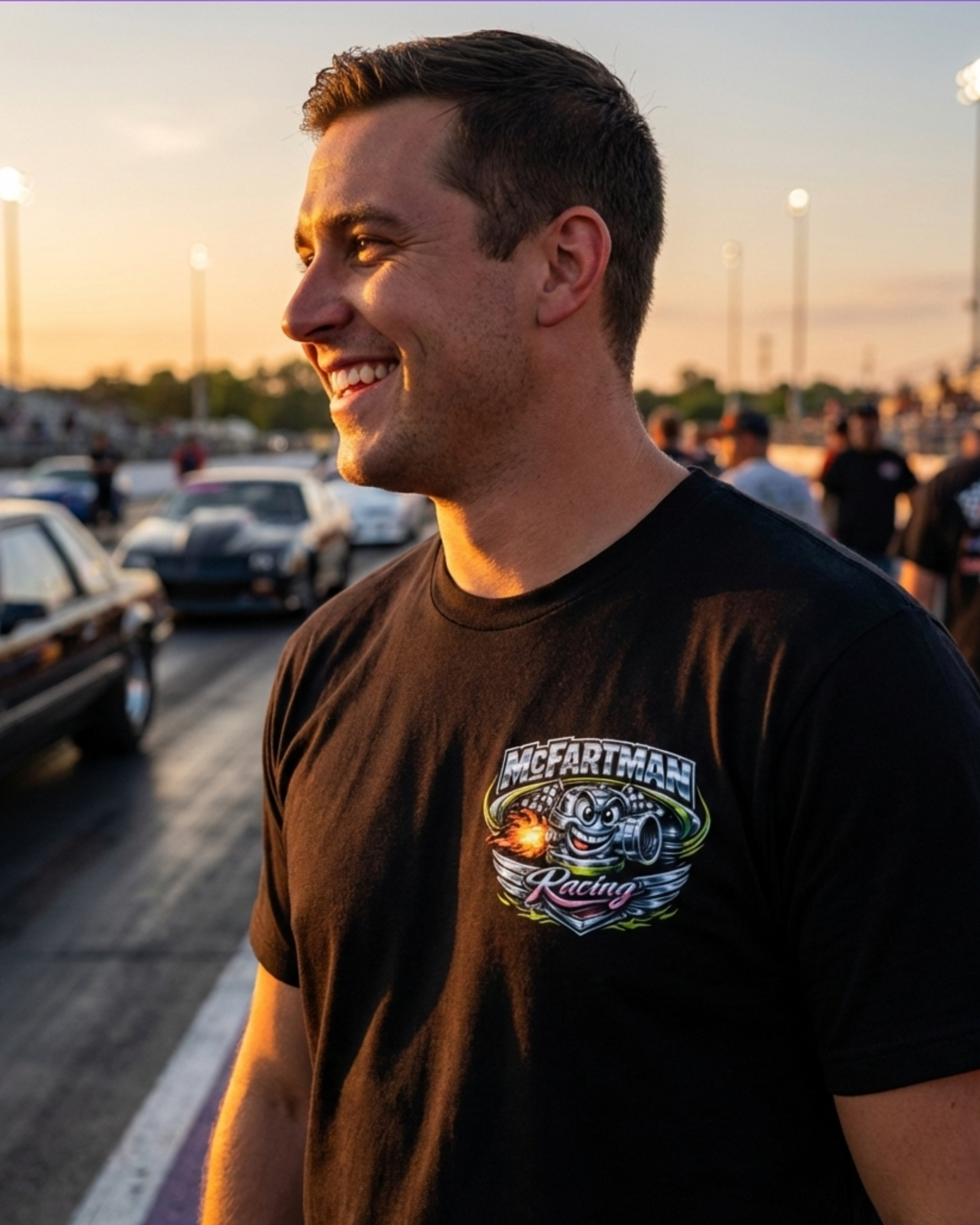 Man wearing a black t-shirt with a McFartman Racing logo at a race track during sunset.