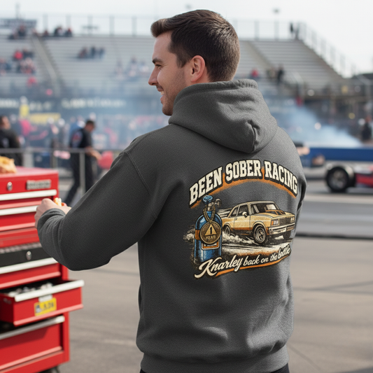 Man wearing a dark gray hoodie with a racing graphic and 'Been Sober Racing Knarley back on the bottle' text at a race track.