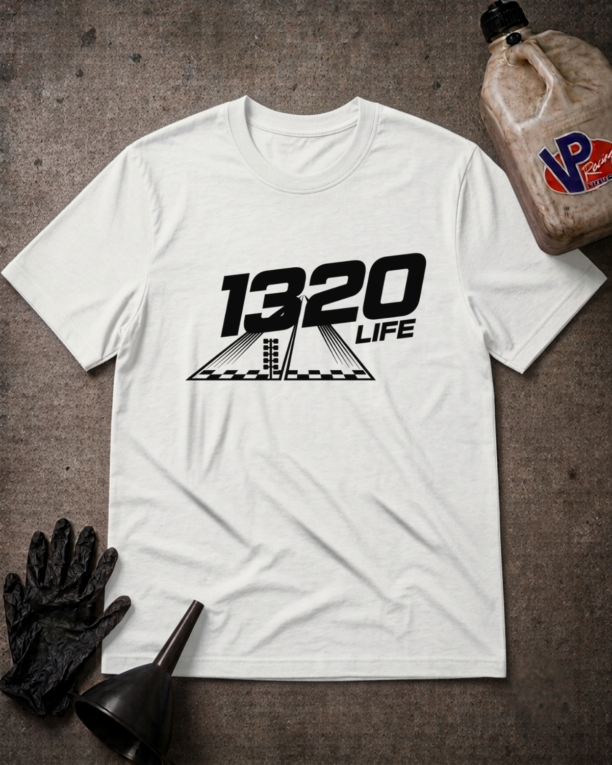 A white short-sleeve graphic T-shirt laid flat on a textured gray surface, featuring a black '1320 LIFE' racing-style print with a checkered flag motif. Black gloves and a funnel are placed near the shirt, and a brown plastic jug with a visible 'VP' logo sits in the upper right.