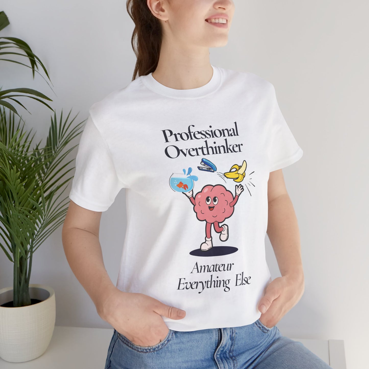 Person wearing a white t-shirt with a graphic and text 'Professional overthinker, Amateur everything else', standing indoors next to a plant.