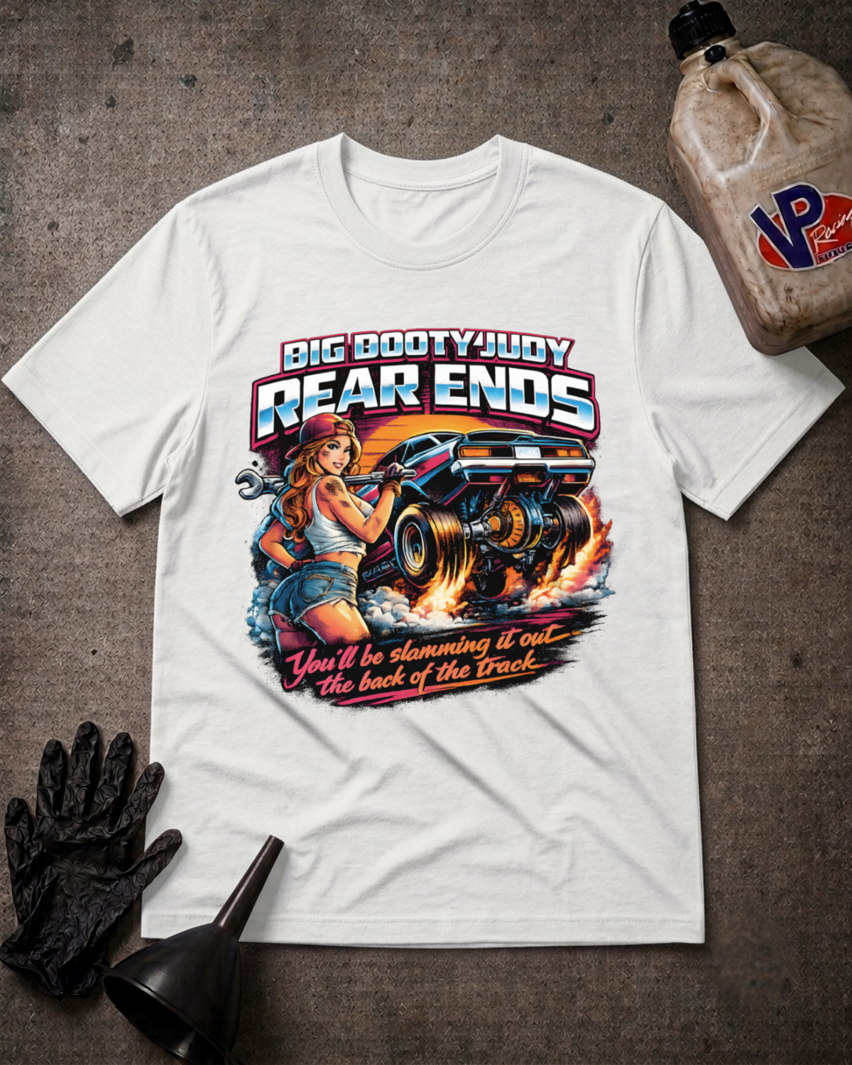 A white graphic t-shirt laid flat on a textured gray surface, surrounded by racing-themed props including a black oil funnel, a black glove, and a plastic jug of VP Racing. The t-shirt features a large graphic of a pin-up girl and a race car with the text 'BIG BOOTYJUDY REAR ENDS' and 'You'll be slamming it out the back of the Track'.