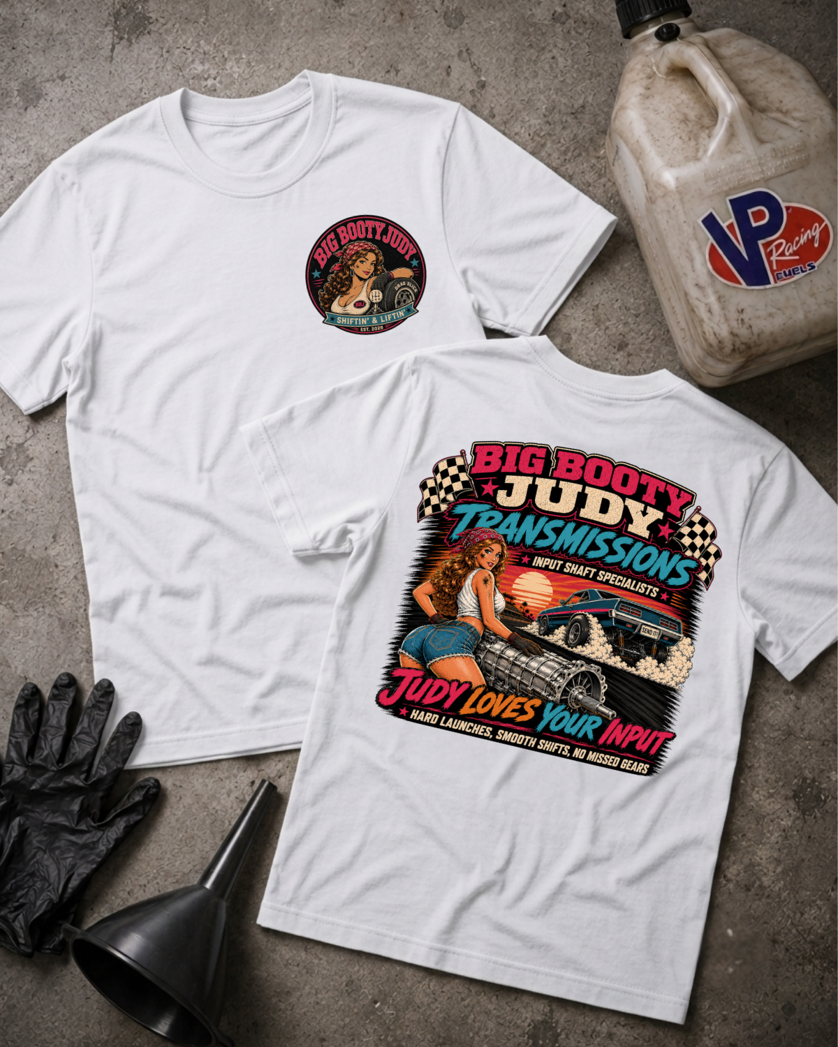 Two white graphic T-shirts laid flat on a gray concrete surface, one showing the front with a small circular chest graphic and the other showing the back with a large colorful automotive-themed print. A plastic jug, black gloves, and a funnel are placed around them as styling props.