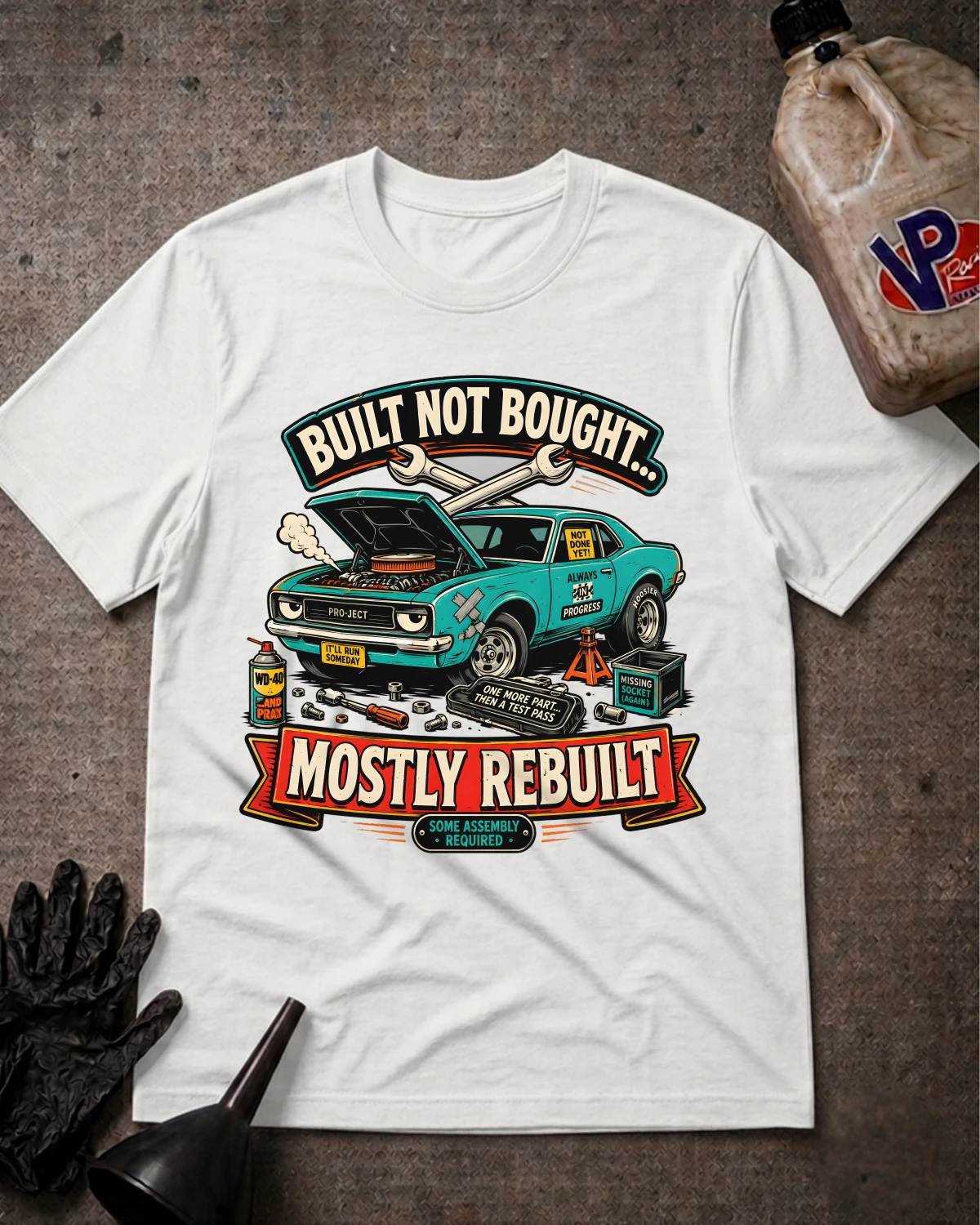 A white graphic t-shirt laid flat on a textured gray surface, featuring a vintage-style teal muscle car with tools and the text 'BUILT NOT BOUGHT.