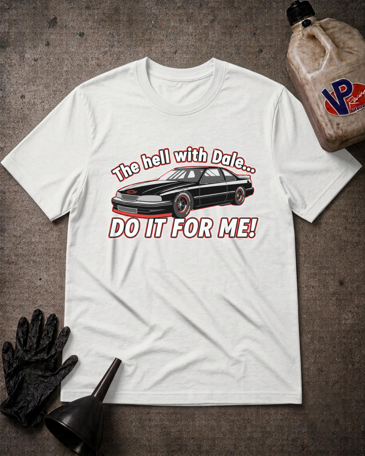 A white graphic t-shirt laid flat on a textured gray surface, featuring a black car and the text 'The hell with Dale Do It For Me.'