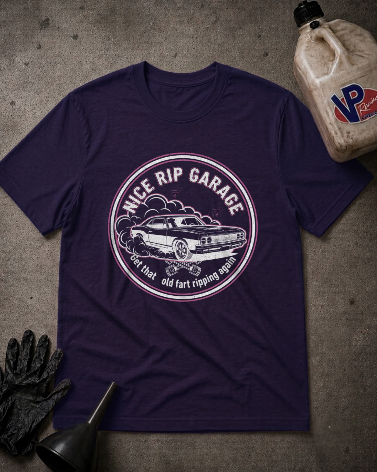 A dark purple crewneck t-shirt with a circular graphic reading 'NICE RIP GARAGE' and 'Get that old fart ripping again' around an illustration of a classic car. The shirt is laid flat on a textured gray concrete surface, styled with a black funnel, black gloves, and a plastic jug of VP Racing fluid in the corners.