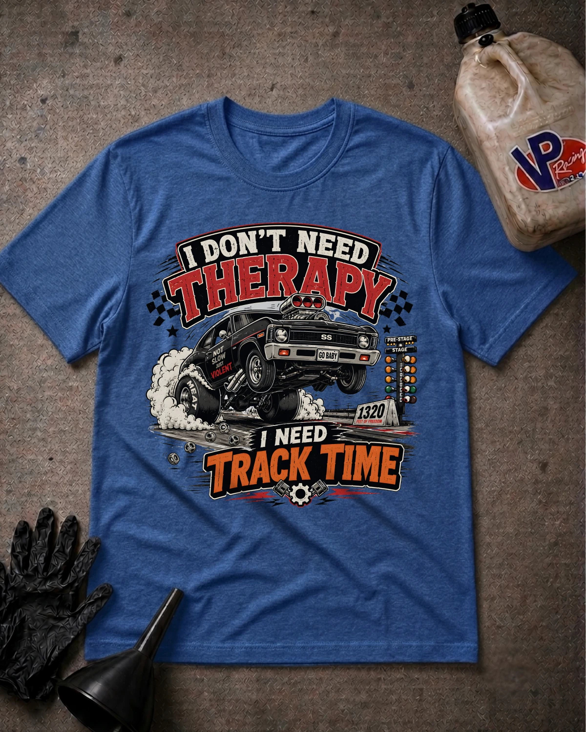 A heather blue graphic t-shirt laid flat on a textured gray surface, surrounded by racing-themed props including a black glove, funnel, and a jug of dirt. The shirt features a graphic of a Decepticon truck with the text 'I DON'T NEED THERAPY' and 'I NEED TRACK TIME'.
