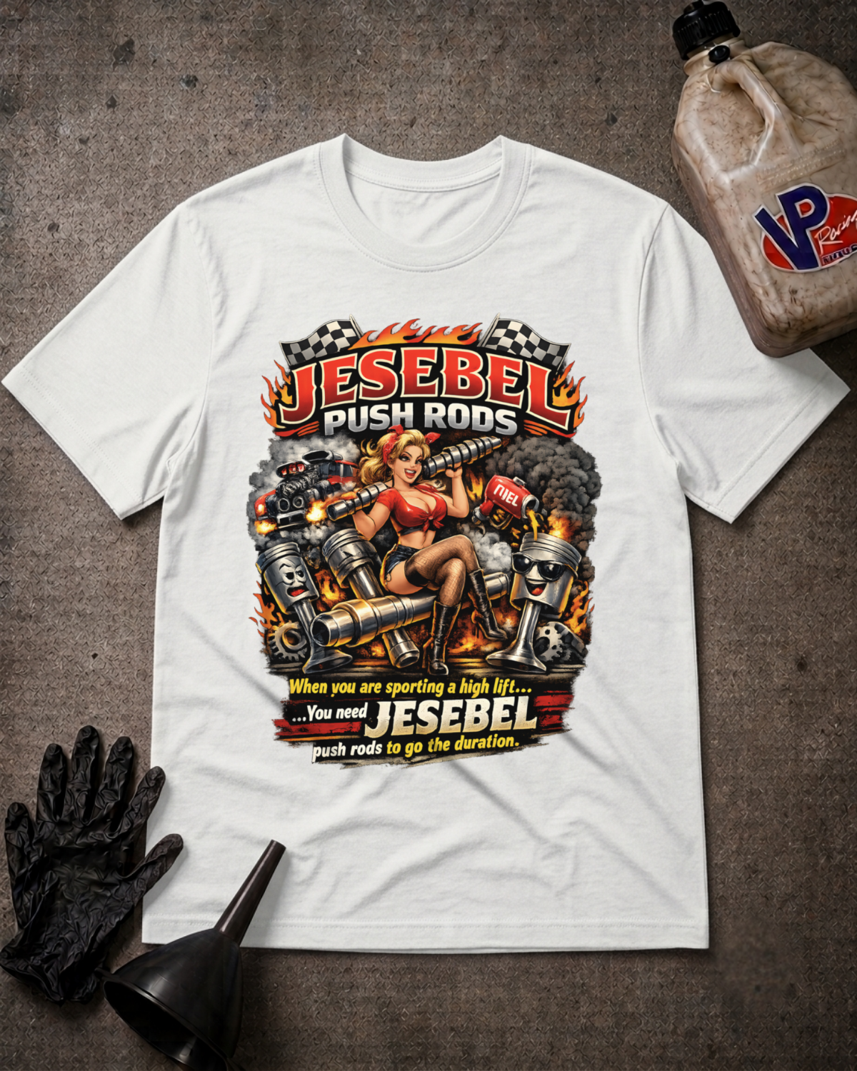 A white short-sleeve graphic T-shirt laid flat on a dark textured surface, surrounded by automotive-themed props including a black oil pan, black gloves, and a plastic jug. The shirt features a large front print with the words 'Jesebel Push Rod' and 'Jesebel push rods to go the duration' over an illustrated pin-up style scene with pistons, flames, and a woman.
