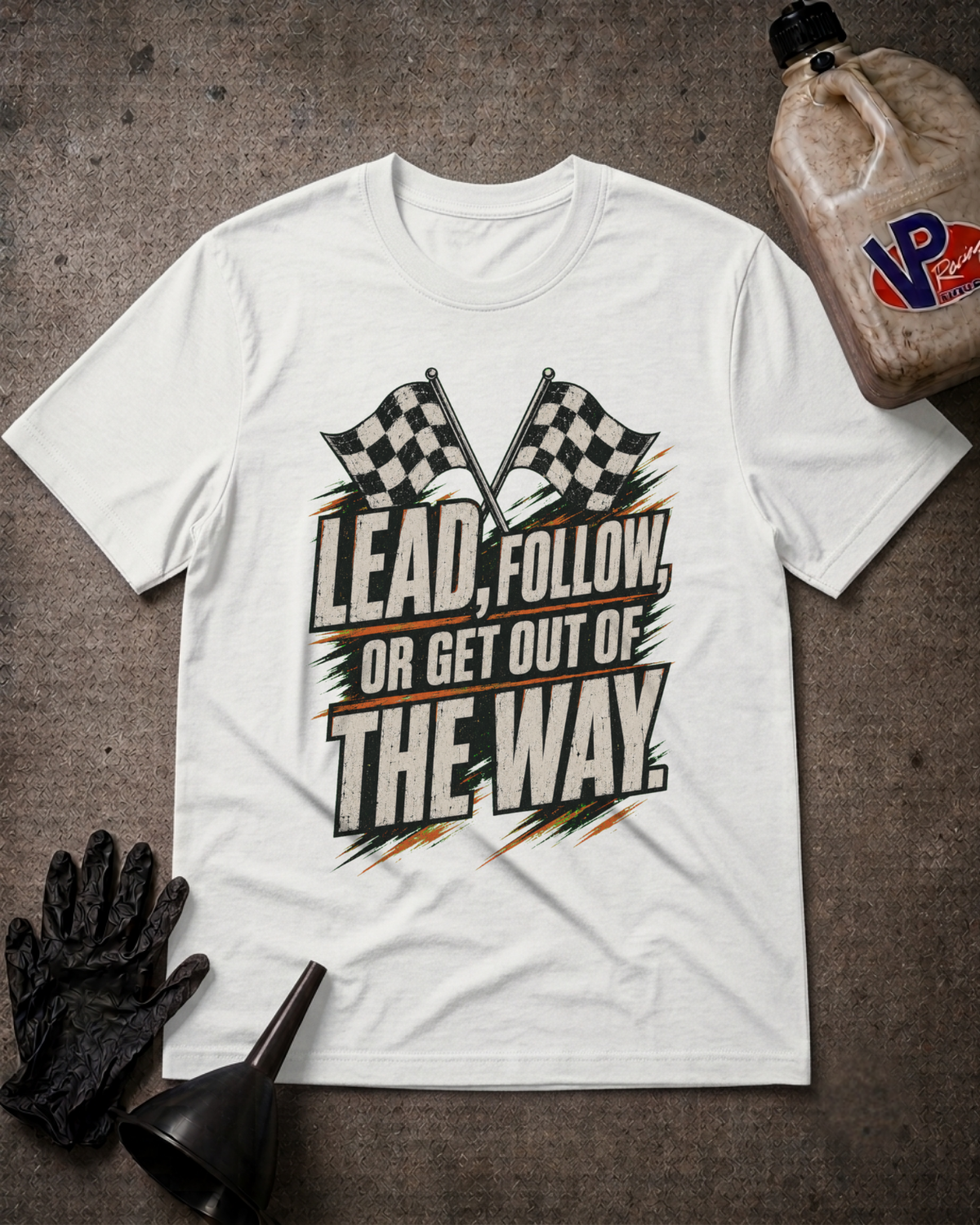 A white graphic t-shirt laid flat on a textured gray surface, surrounded by racing-themed props including a black glove, funnel, and a jug of VP Racing fluid. The shirt features a bold graphic with two checkered flags and the text 'LEAD, FOLLOW, OR GET OUT OF THE WAY.