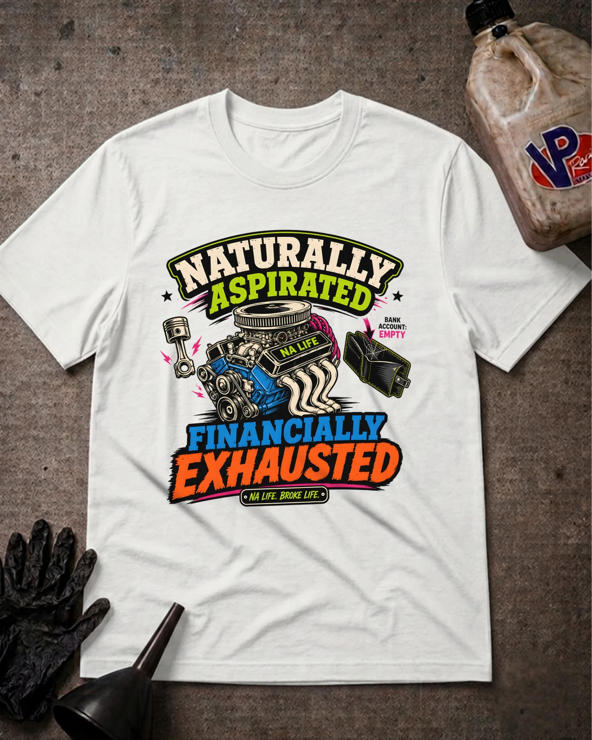 A white graphic t-shirt laid flat on a dark textured surface, featuring a colorful engine illustration and the text 'Naturally Aspirated FINANCALLY EXHAUSTED'. A plastic jug, black gloves, and a funnel are styled around the shirt.