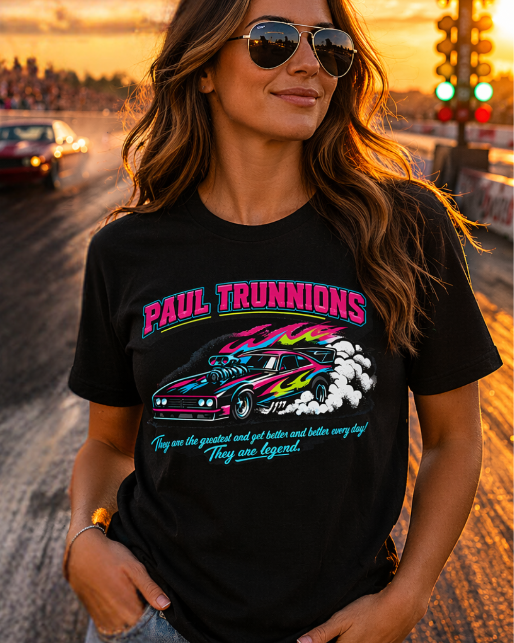 Woman wearing a black t-shirt with colorful graphics and 'Paul Trunnions They are the greatest and get better and better every day! They are lengend' text, standing on a drag strip at sunset.