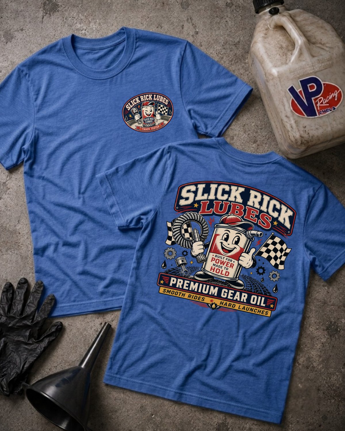 Two blue graphic T-shirts laid flat on a textured gray surface, with a black oil jug, black gloves, and a funnel nearby. The front shirt shows a small circular 'Black Rick Lube' logo on the left chest, and the back shirt shows a large 'Slick Rick Lubes Premium Gear Oil' graphic with an illustrated oil can and racing elements.
