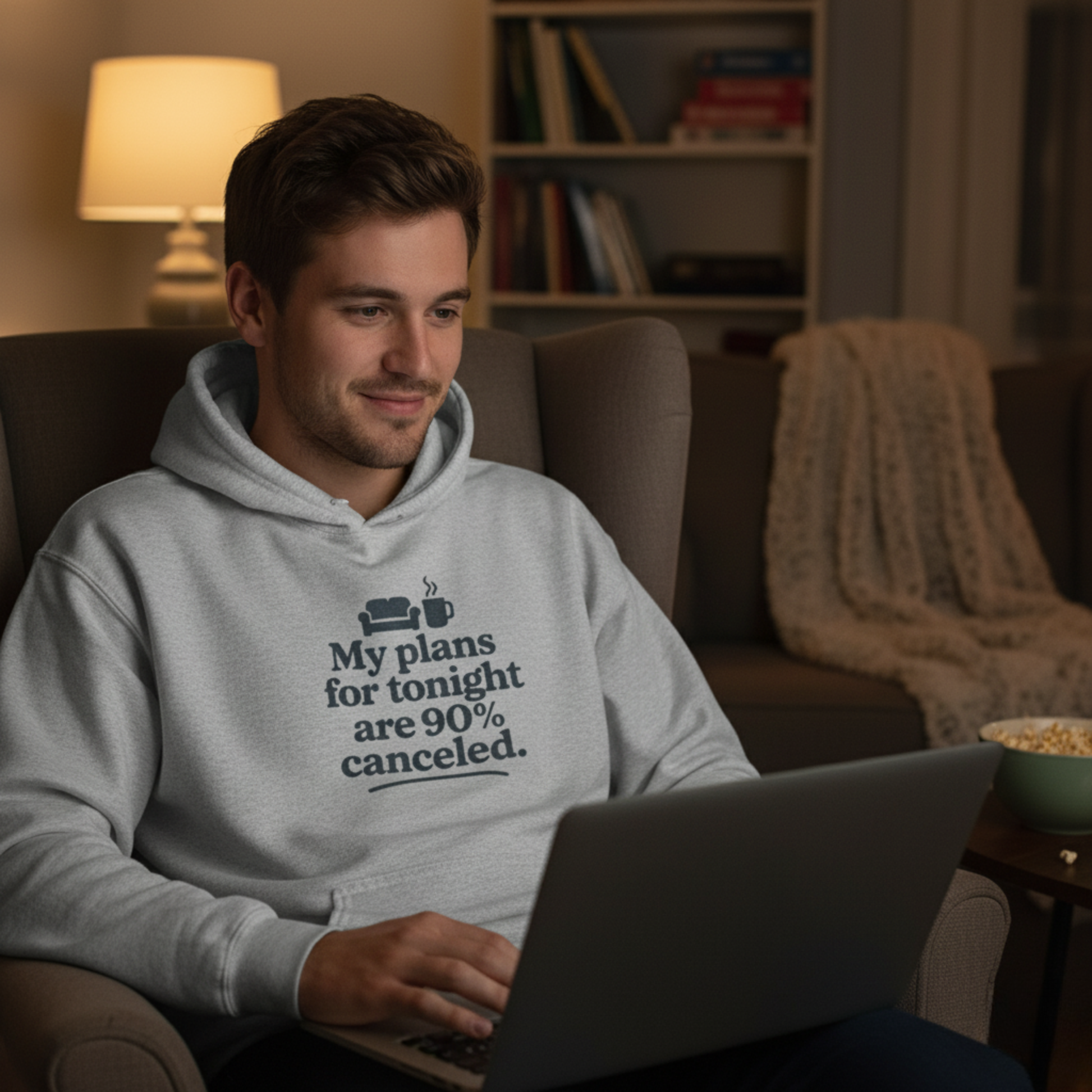 Man sitting in a chair using a laptop with a hoodie on with text 'My plans for tonight are 90% canceled', in a cozy living room setting.