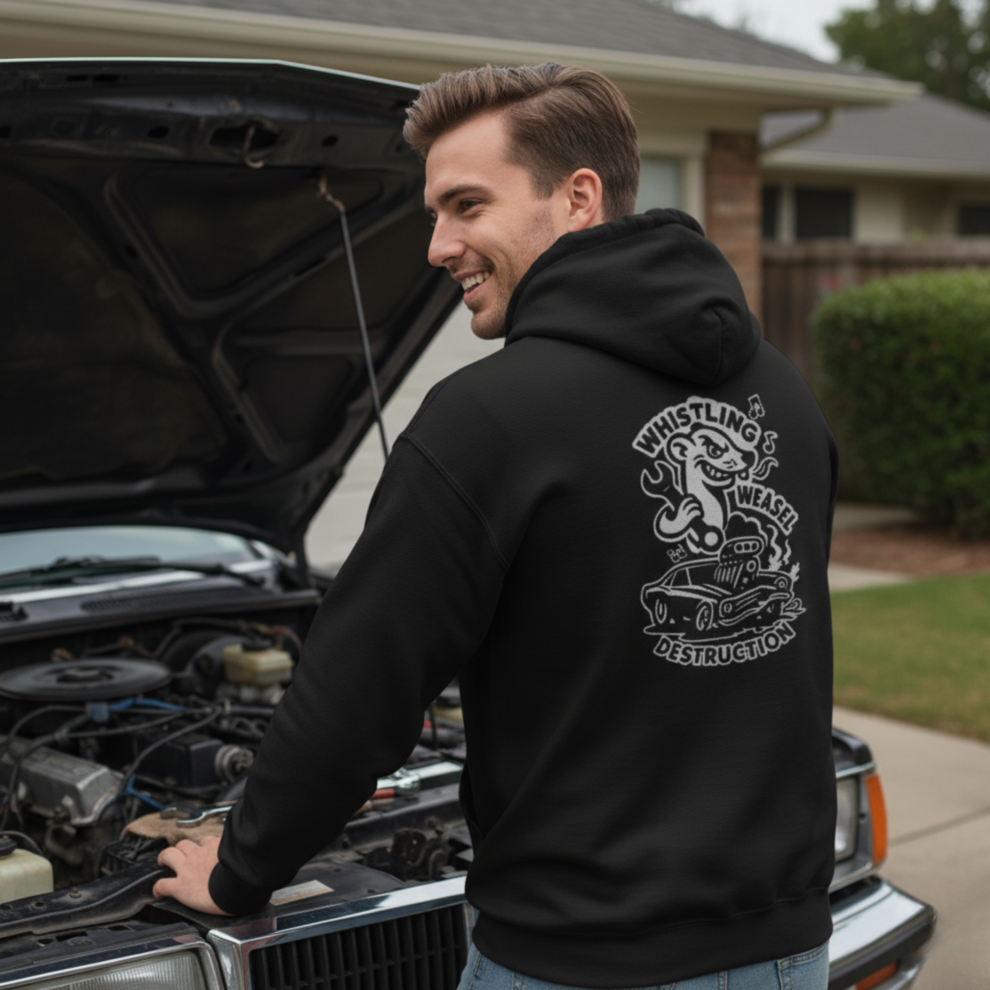 Person wearing black Gildan 18500 hoodie with large white back graphic featuring cartoon weasel and flame-spitting car. Standing beside open car hood in residential garage setting. Ideal for drag racing fans and motorsport lifestyle.