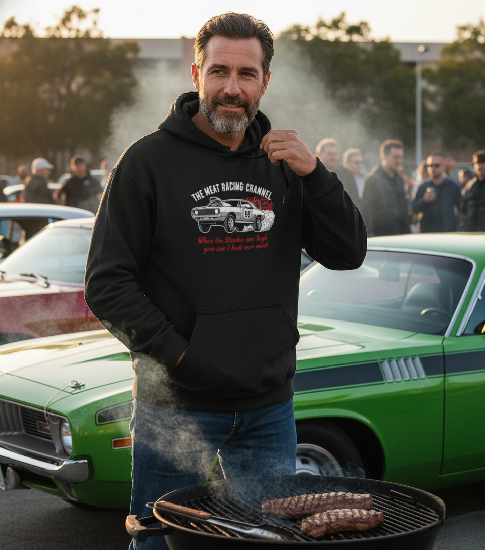 Man wearing black The Meat Racing Channel hoodie, grilling steaks next to a green classic muscle car at sunset.