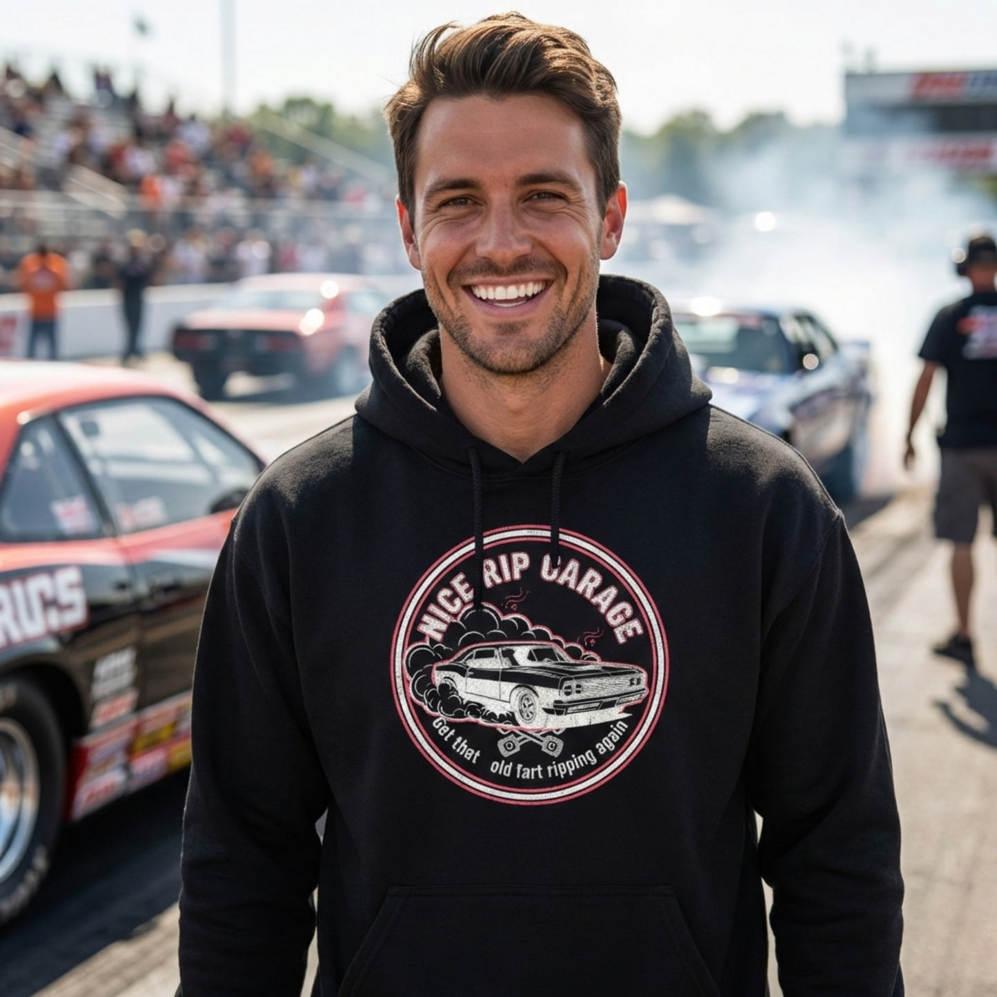 Man wearing a black hoodie with a graphic design and 'Nice Rip Garage Get that old fart ripping again' text at a race track