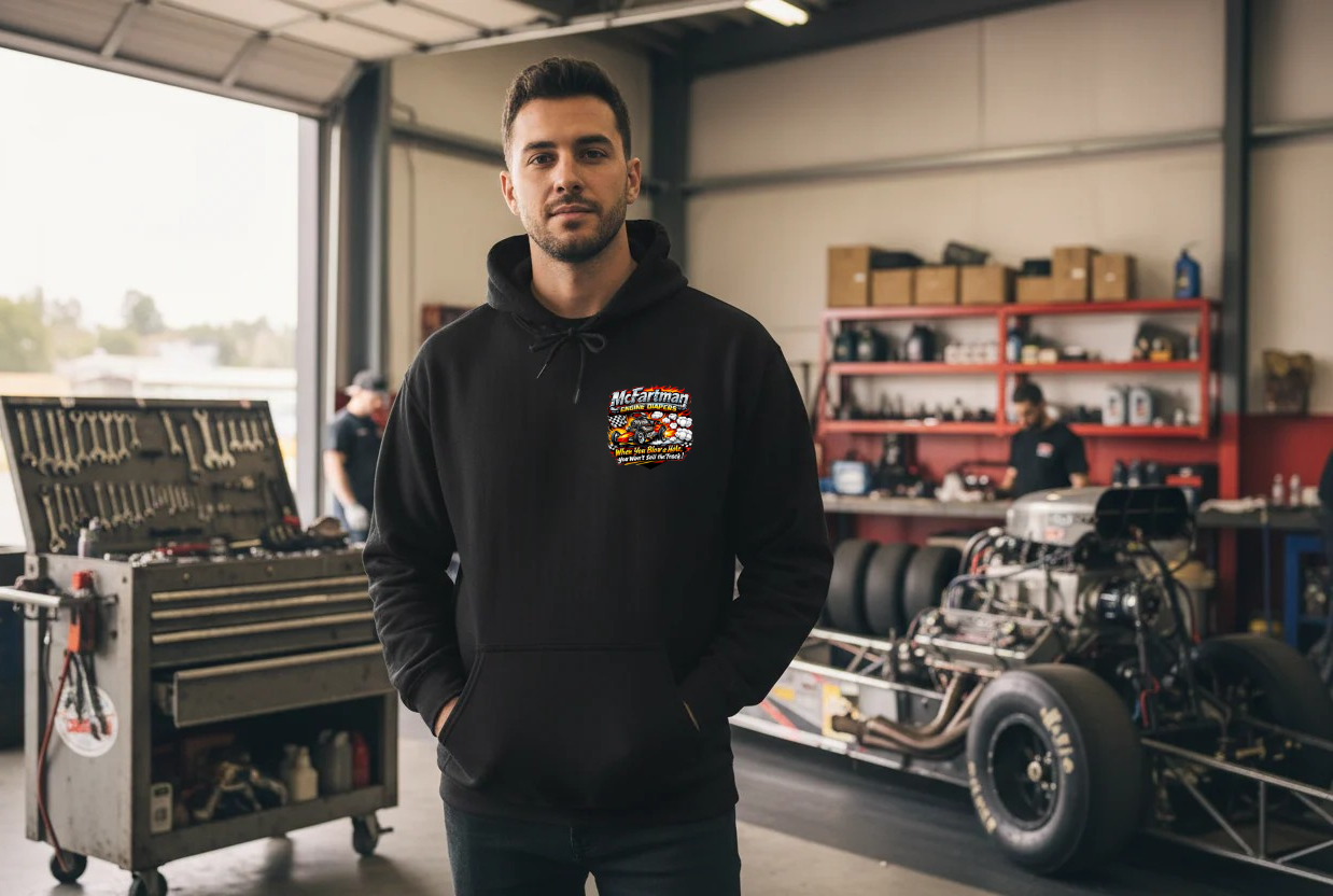 Man wearing a black hoodie with a 'McFartman Engine Diapers' logo in a garage with automotive equipment and a car engine.