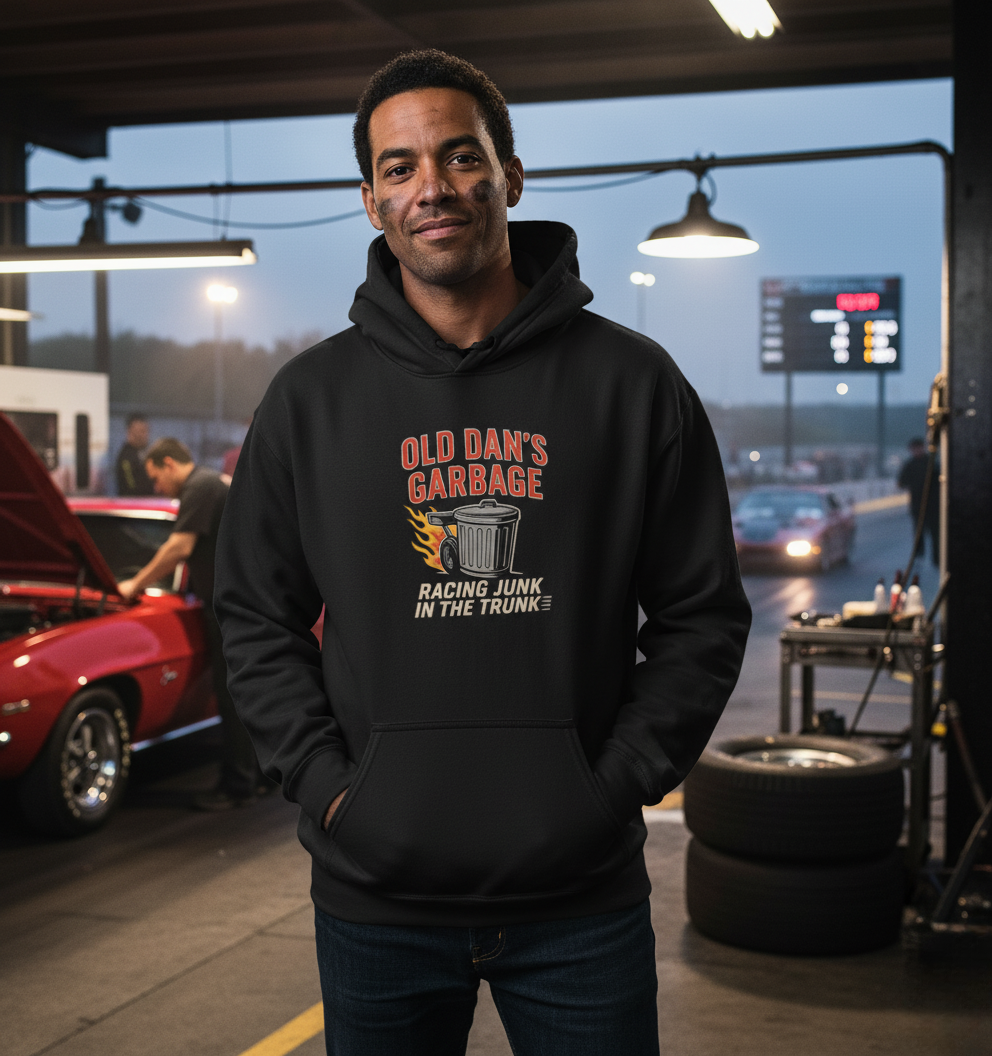 Man wearing black Old Dan's Garbage racing hoodie in a drag strip pit lane with a red muscle car.