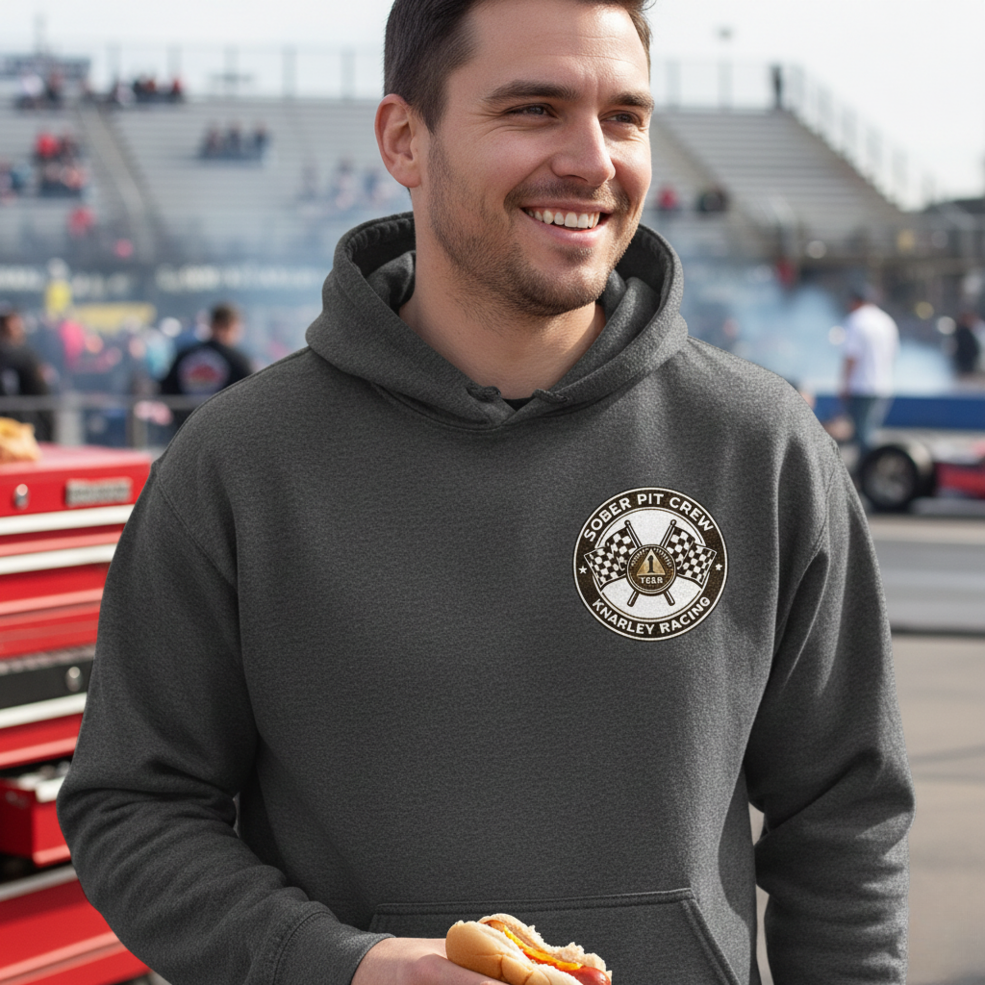 Man wearing a dark gray hoodie with a 'Sober Pit Crew Knarley Racing' logo, standing in a pit lane.