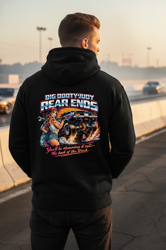 Person wearing a black hoodie with a colorful graphic and 'Big Booty Judy Rear Ends You'll be slamming it out the back of the track' text on the back, standing on a race track with vehicles in the background.