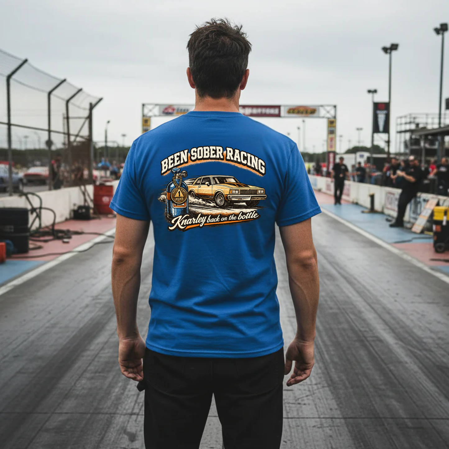 Person wearing a blue t-shirt with racing graphics and 'Been Sober Racing Knarley back on the bottle' text at a race track.