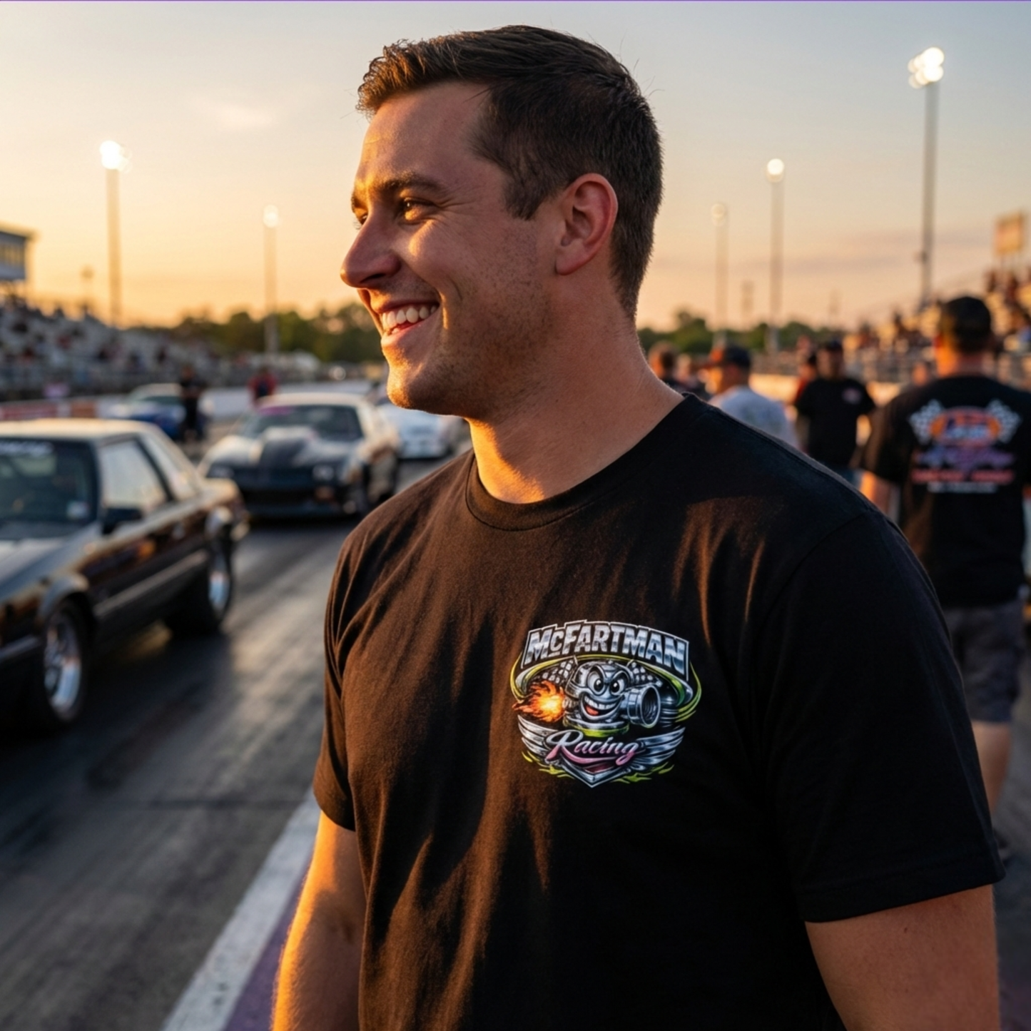 Man wearing a black t-shirt with a McFartman Racing logo at a race track during sunset.