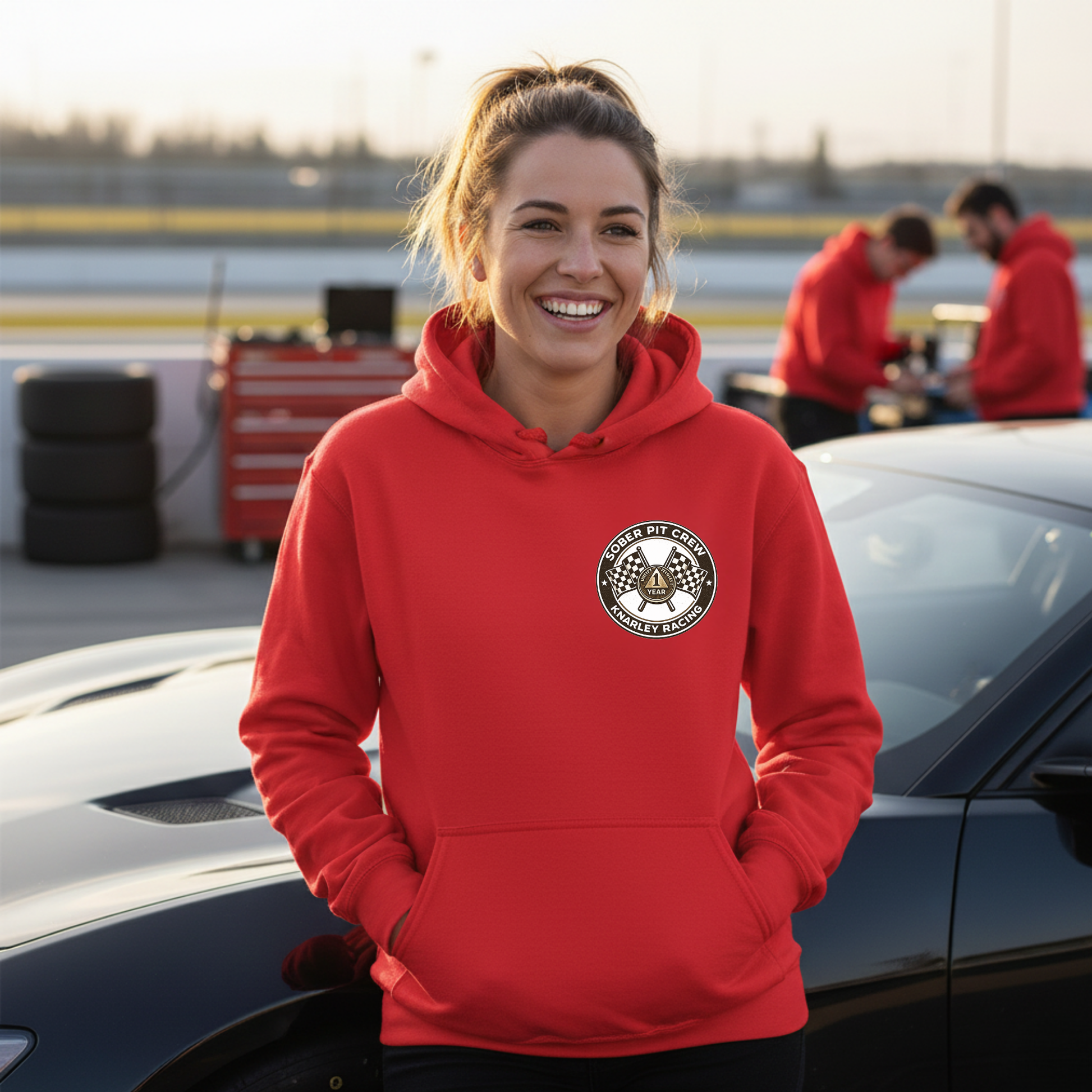 Woman wearing a red hoodie with a 'Sober Pit Crew Knarley Racing' logo, standing next to a car on a racetrack.