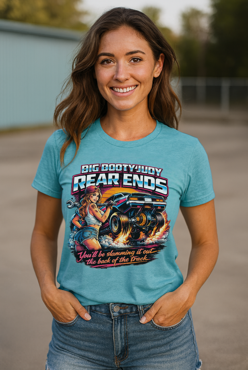 Woman wearing a blue t-shirt with a graphic design of a woman and a car and 'Big Booty Judy Rear Ends You'll be slamming it out the back of the track' text, standing outdoors.