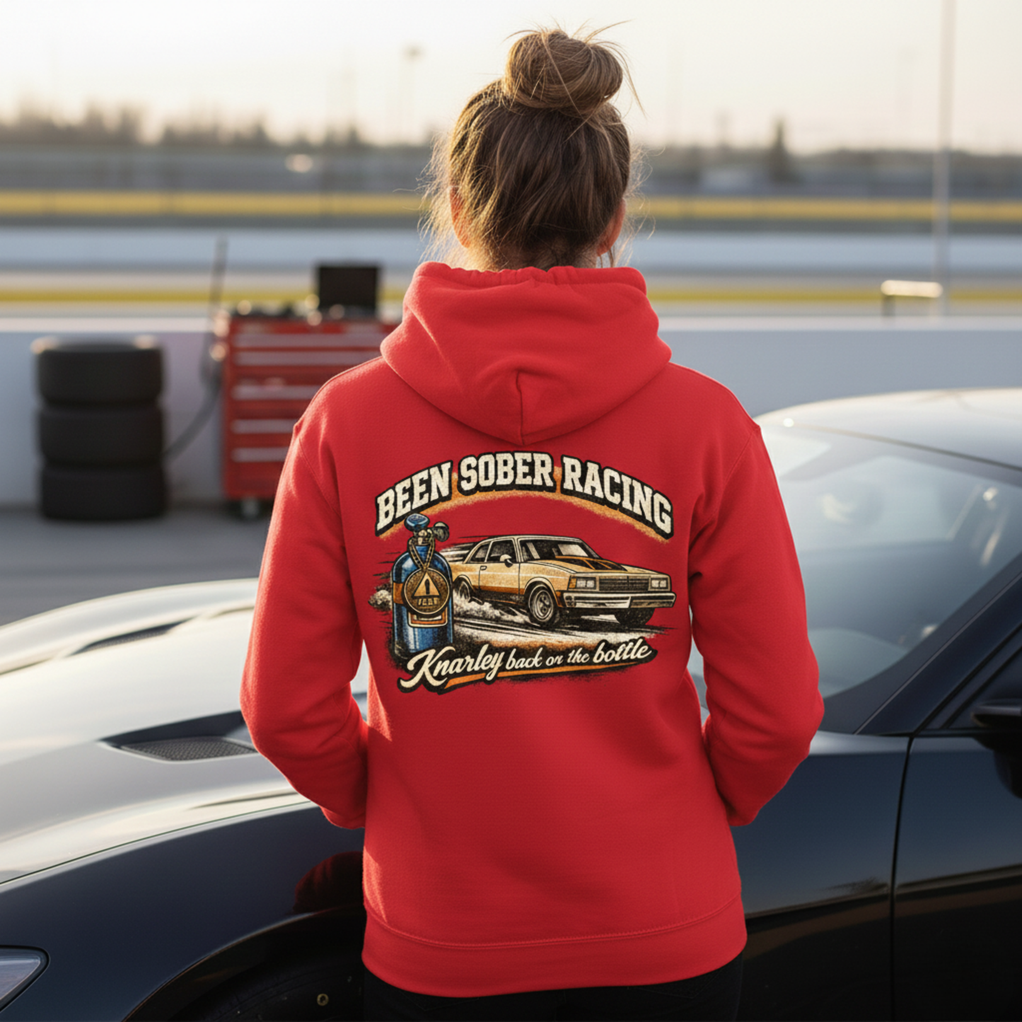 Person wearing a red hoodie with 'Been Sober Racing' text and graphic, standing next to a car on a racetrack.