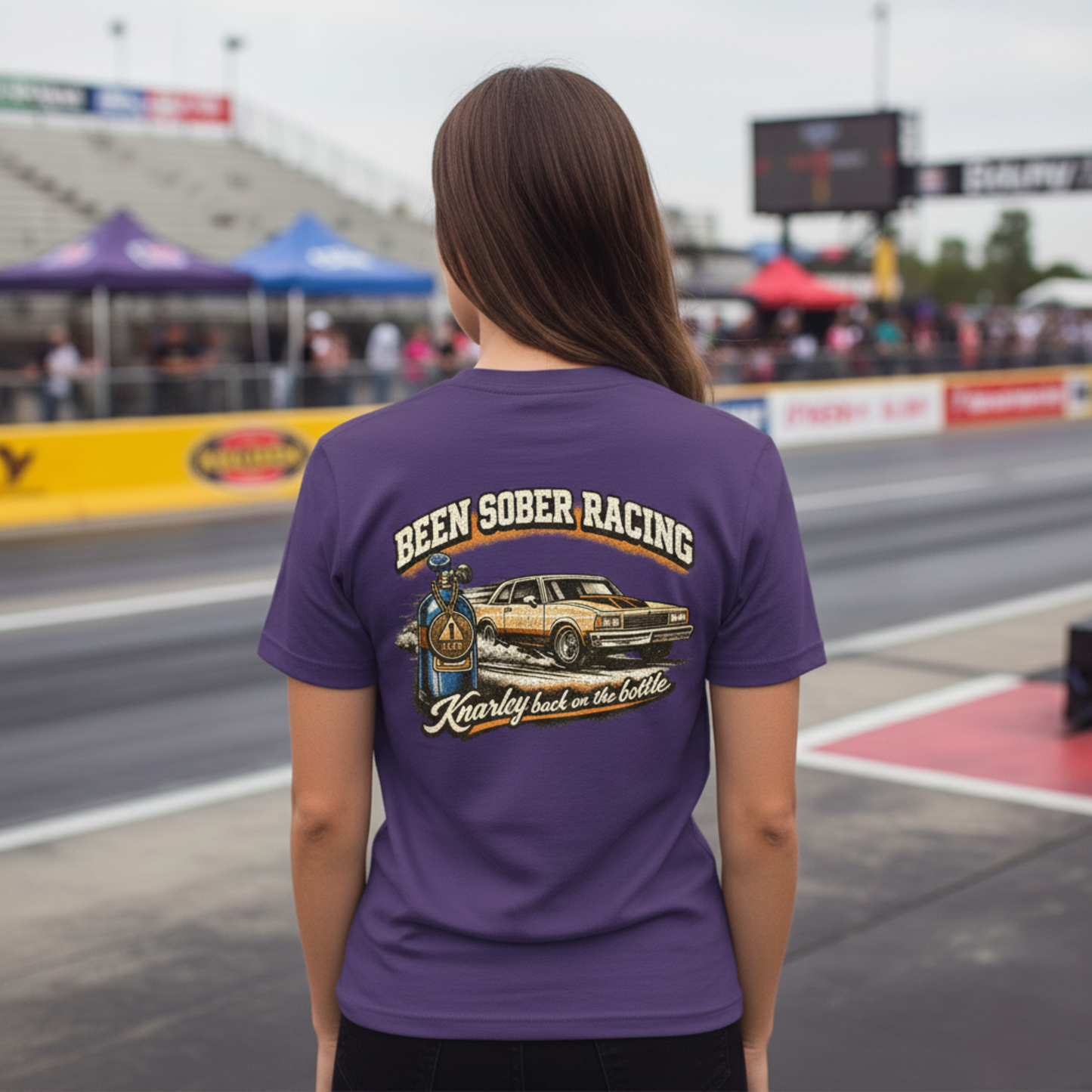 Person wearing a purple t-shirt with a graphic and 'Been Sober Racing Knarley back on the bottle' text at a race track.
