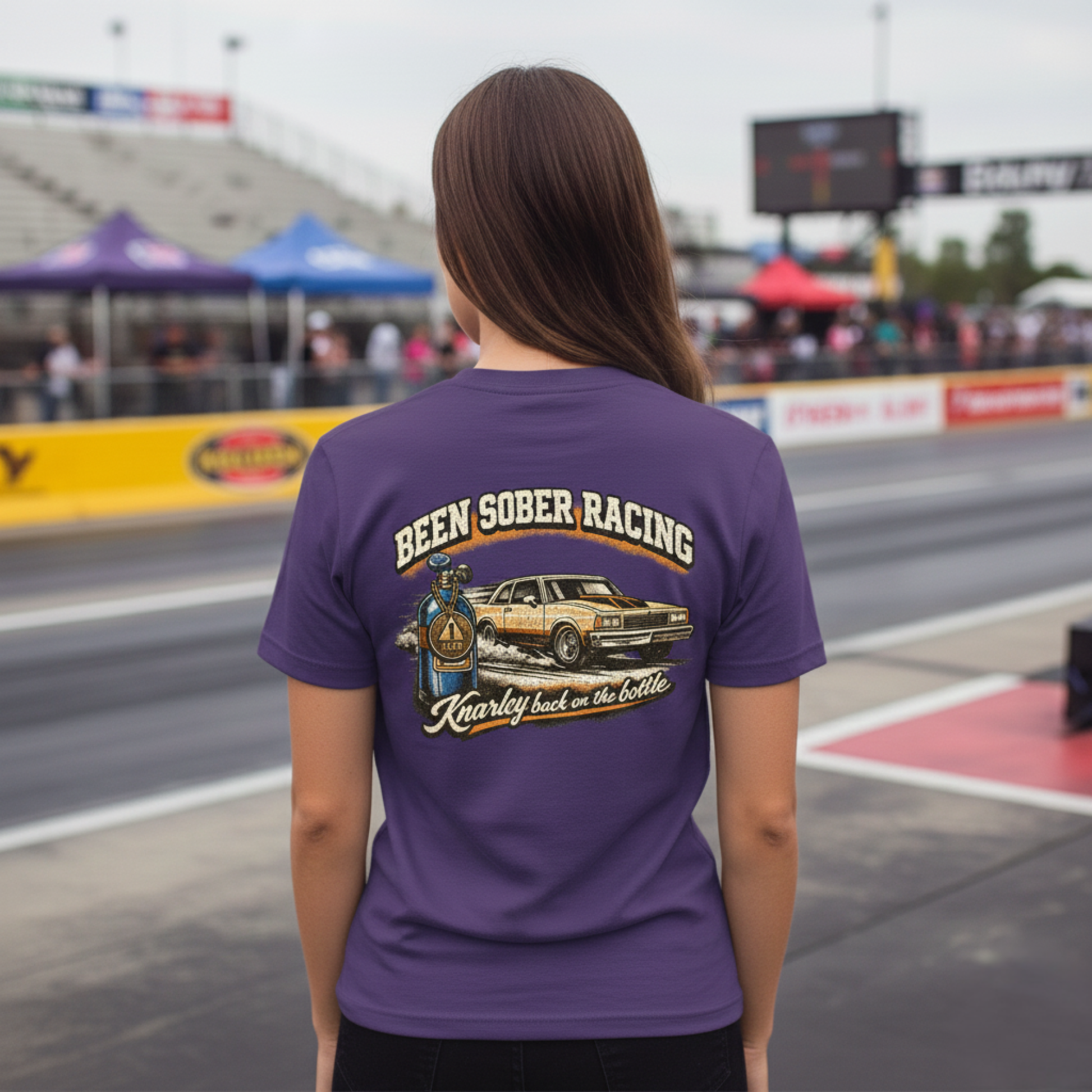 Person wearing a purple t-shirt with a graphic and 'Been Sober Racing Knarley back on the bottle' text at a race track.