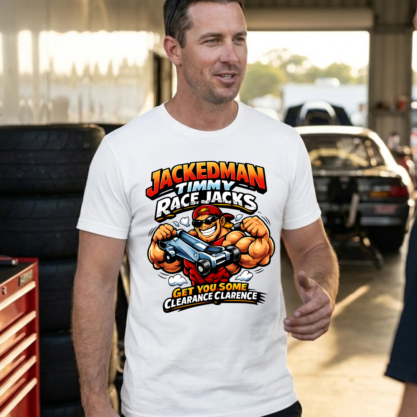 Man wearing a white t-shirt with a graphic design 'Jackedman Timmy Race Jacks Get You Some Clearance Clarence' in an outdoor setting with cars and tires.
