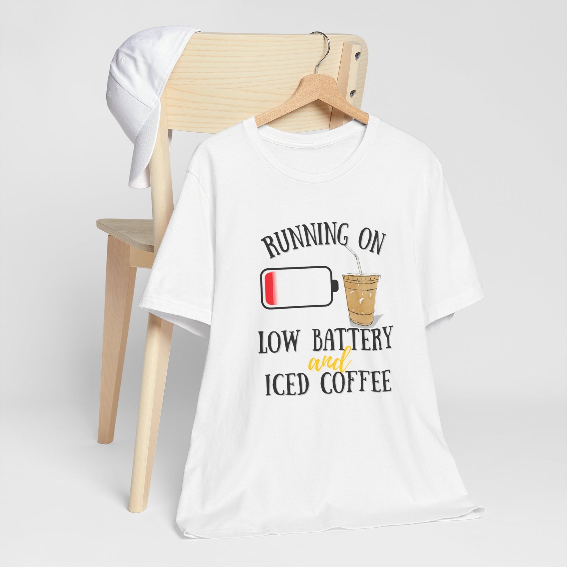 White t-shirt with humorous text 'Running on low battery and iced coffee' design on a wooden chair against a white background