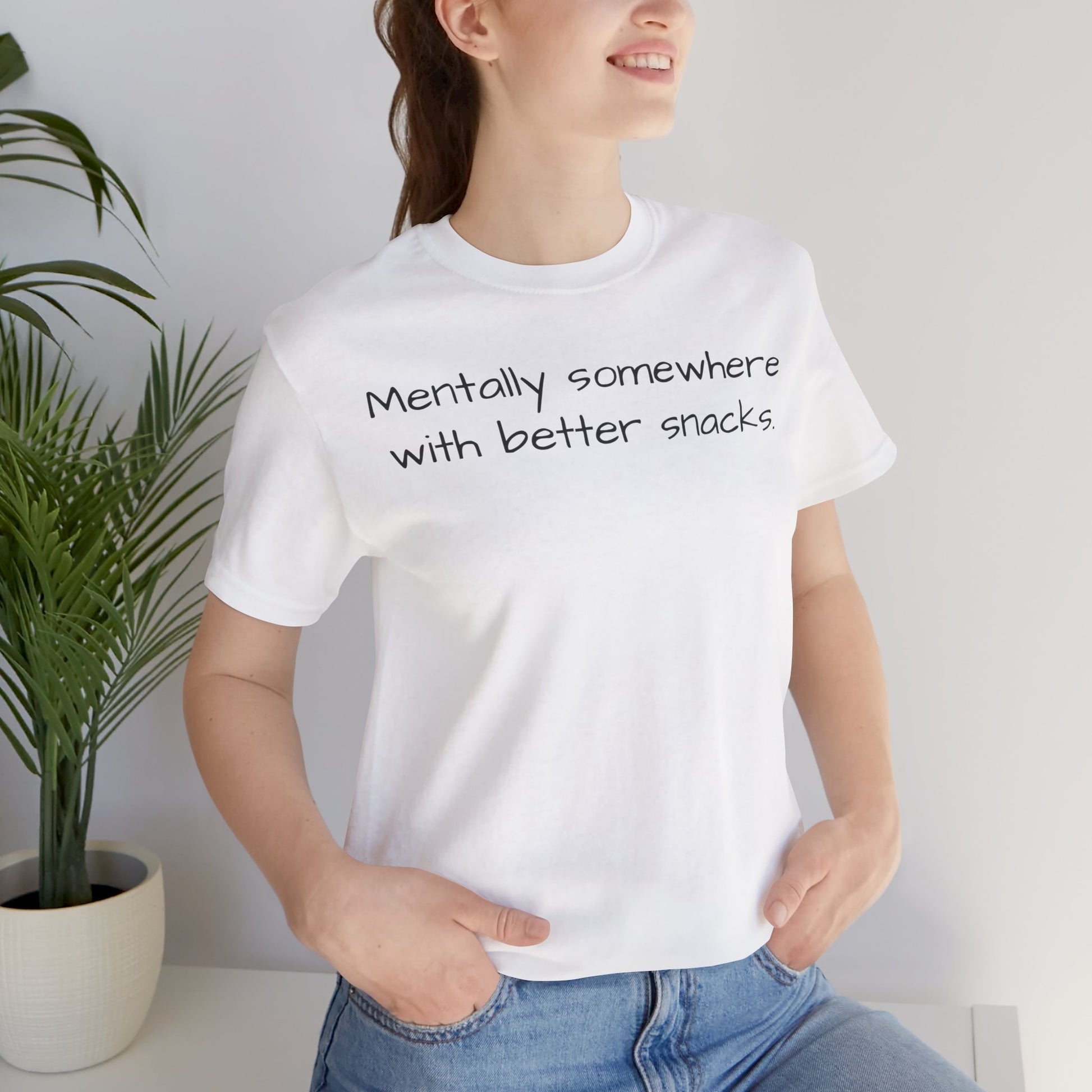 Person wearing a white t-shirt with text 'Mentally somewhere with better snacks', standing next to a plant indoors.