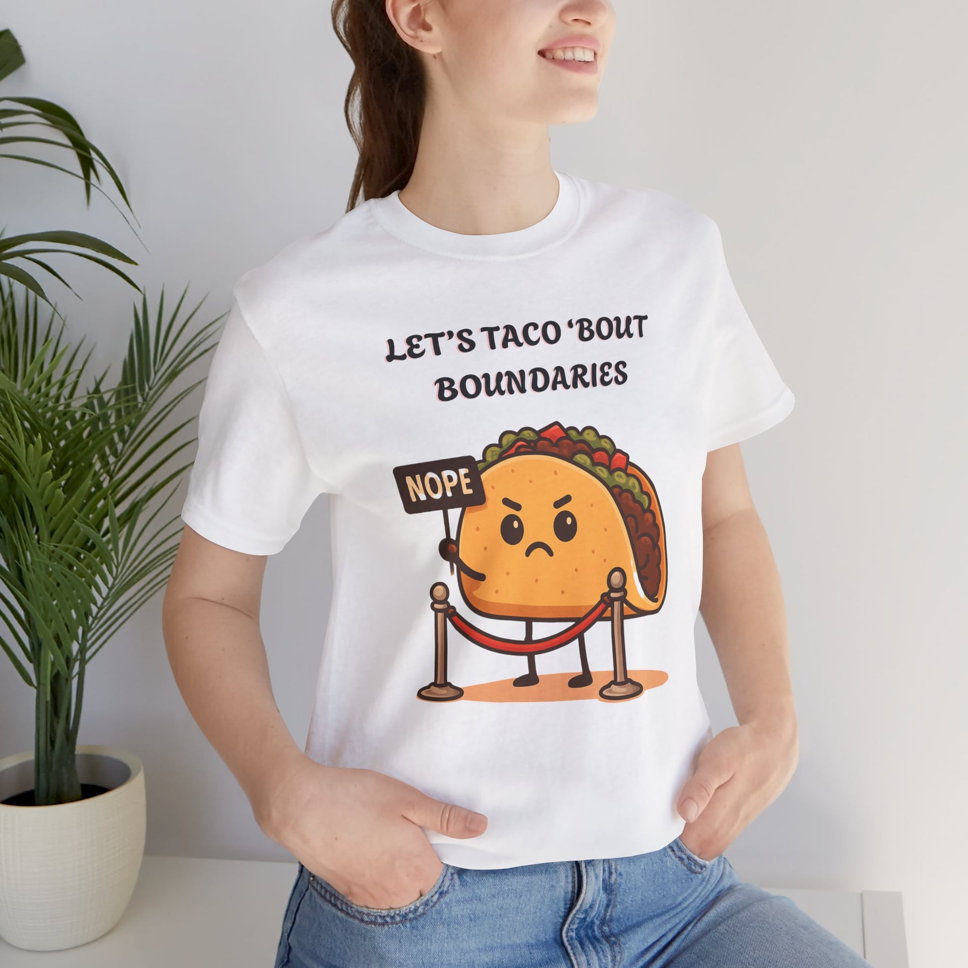 Person wearing a white t-shirt with a taco graphic and text 'Let's taco 'bout boundaries', standing indoors next to a plant.