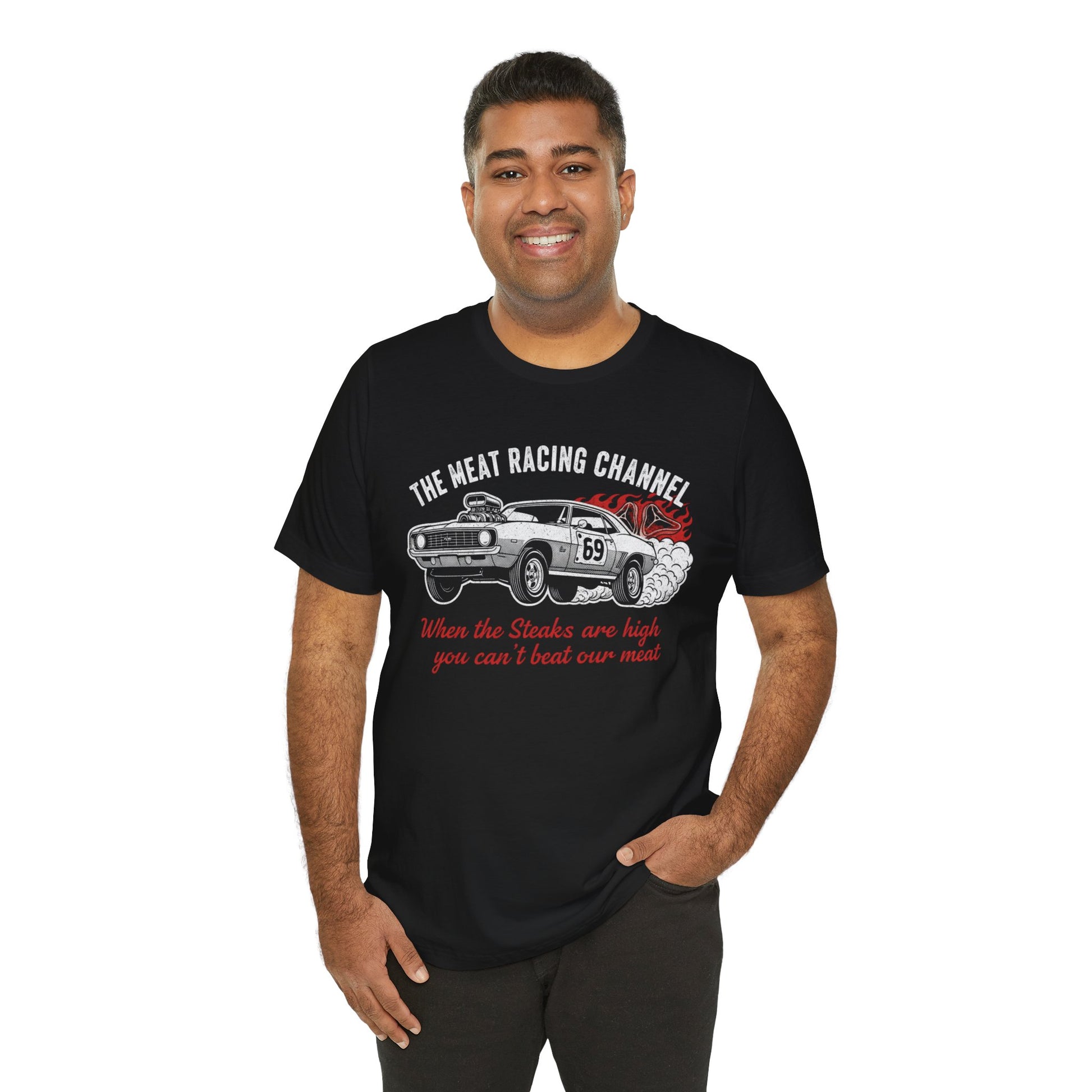 Person wearing a black Bella+Canvas 3001 T‑shirt featuring a bold drag racing graphic. The design shows a vintage muscle car with the number 69, white smoke and red flames, the distressed white title “The Meat Racing Channel” above, and the red script tagline “When the Steaks are high you can’t beat our meat” below. Funny drag racing parody shirt with retro hot rod style, perfect for car enthusiasts, gearheads, and humor‑driven racing apparel fans.