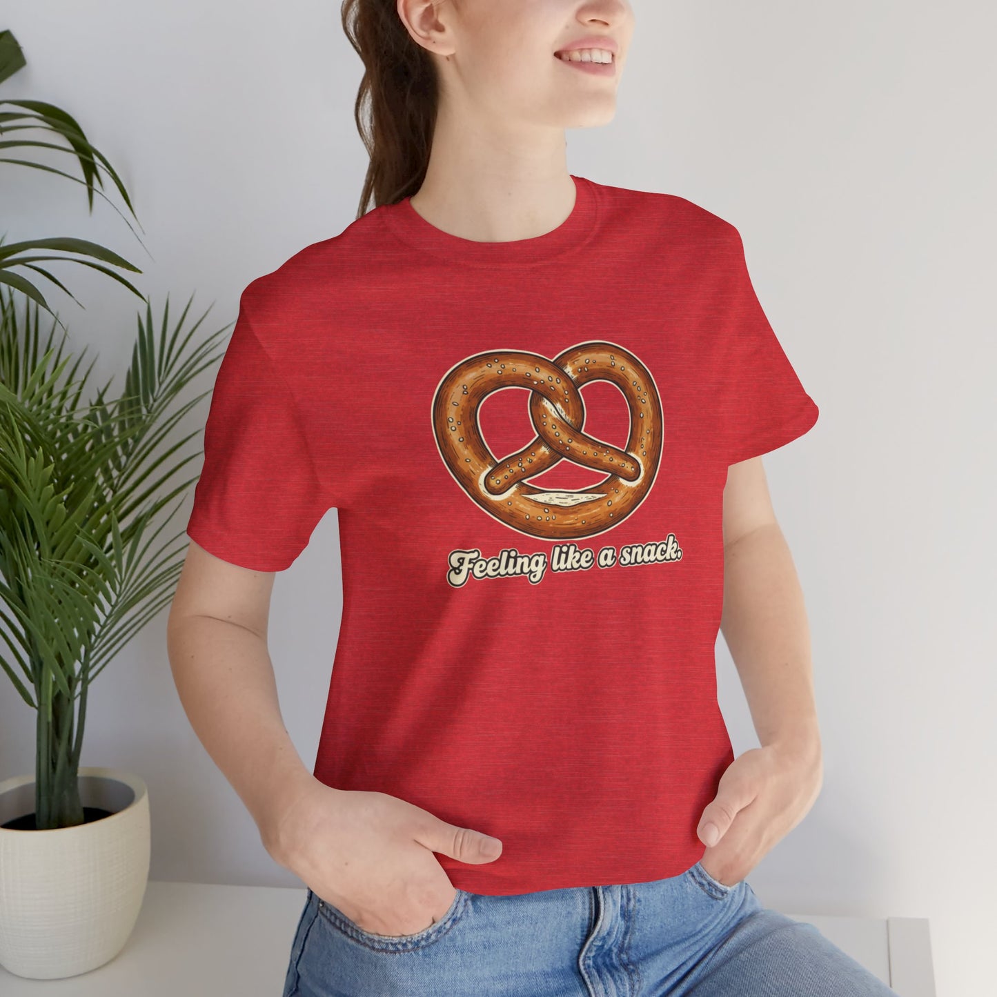 Person wearing a red t-shirt with a pretzel graphic and text 'Feeling like a snack', standing indoors next to a plant.