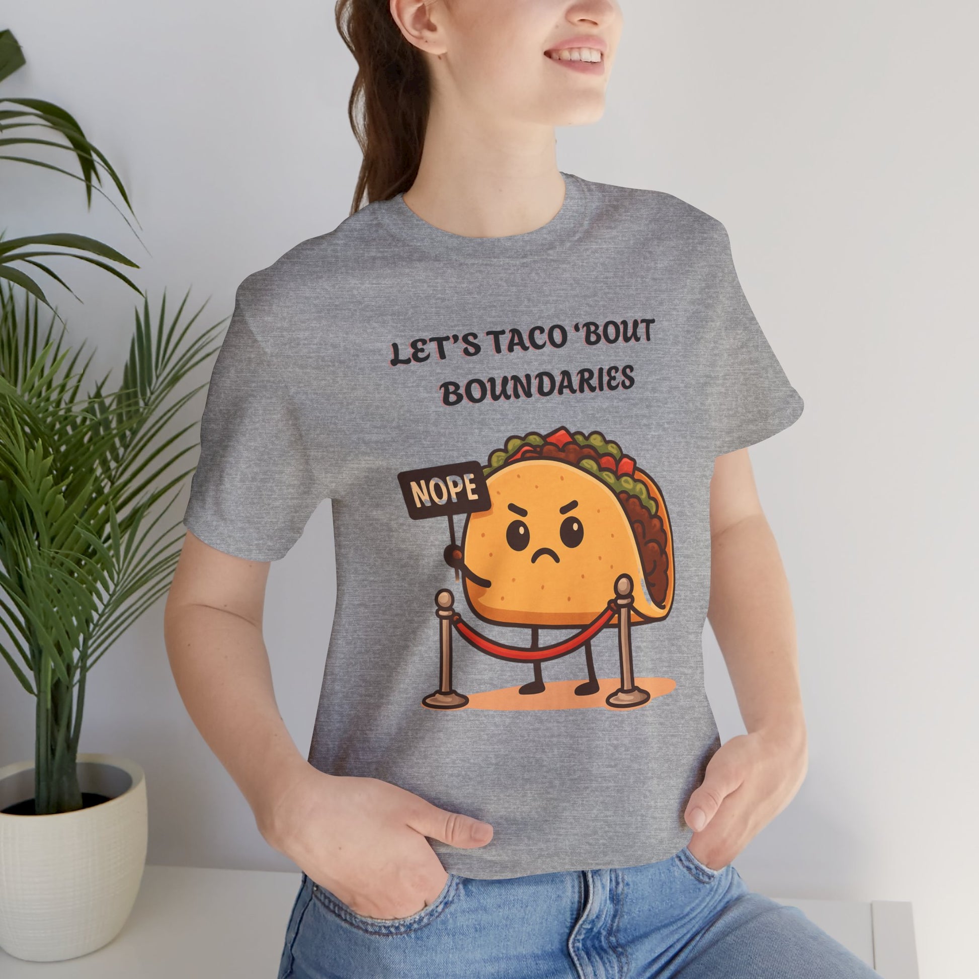Person wearing a gray t-shirt with a taco graphic and text 'Let's taco 'bout boundaries', standing indoors next to a plant.