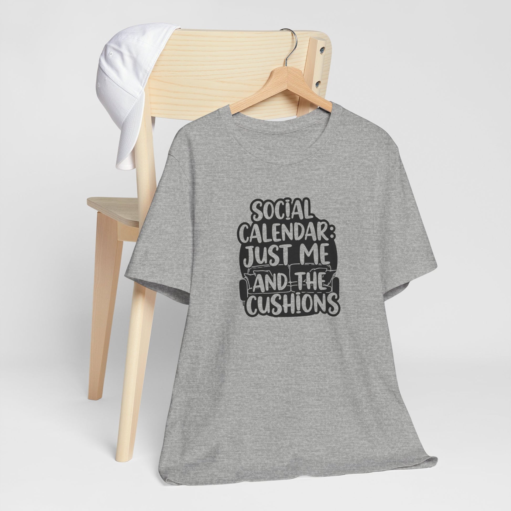 Gray t-shirt with text 'Social Calendar: Just Me and the Cushions' on a wooden chair.
