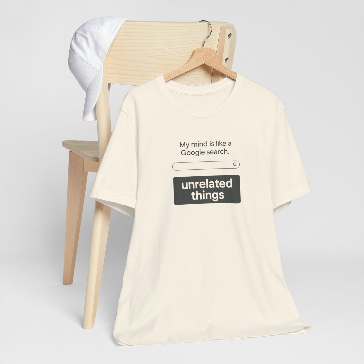 Beige t-shirt with humorous text design on a wooden chair against a light gray background. T-shirt has a search bar graphic with text above that says, 'My mind is like a Google search', and text below that says, 'unrelated things.'