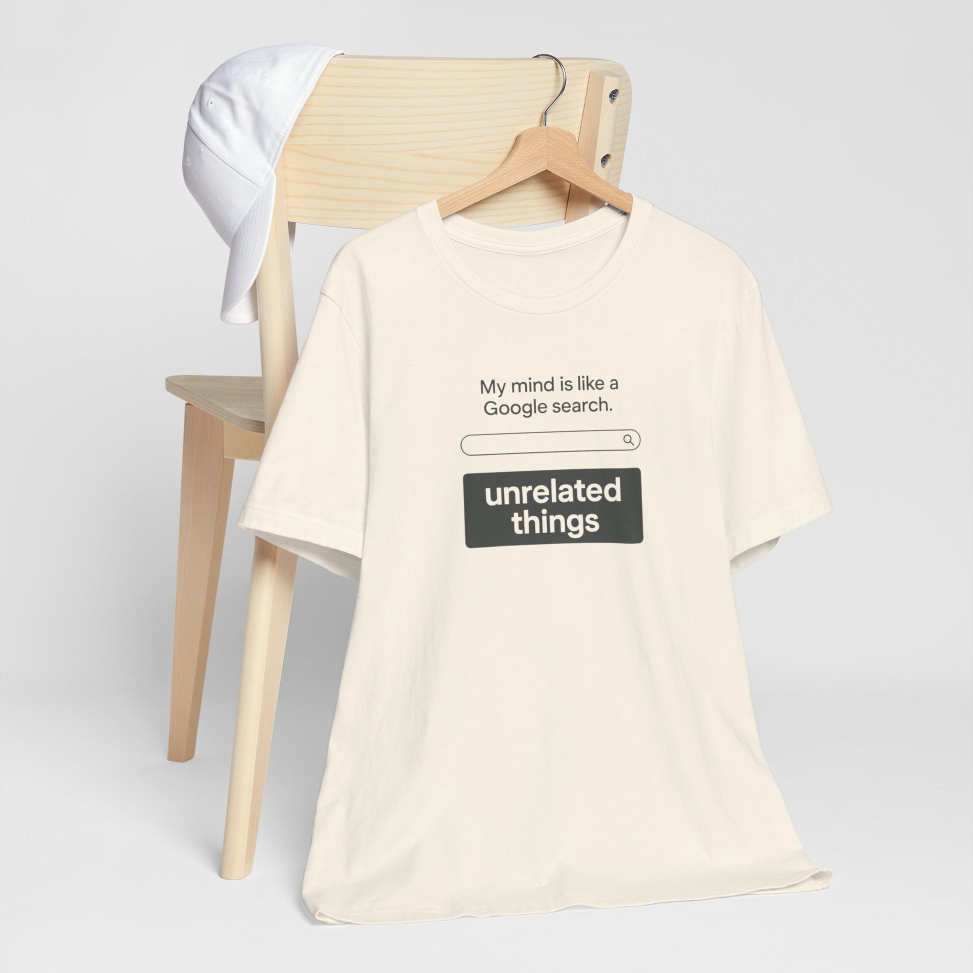 Beige t-shirt with humorous text design on a wooden chair against a light gray background. T-shirt has a search bar graphic with text above that says, 'My mind is like a Google search', and text below that says, 'unrelated things.'