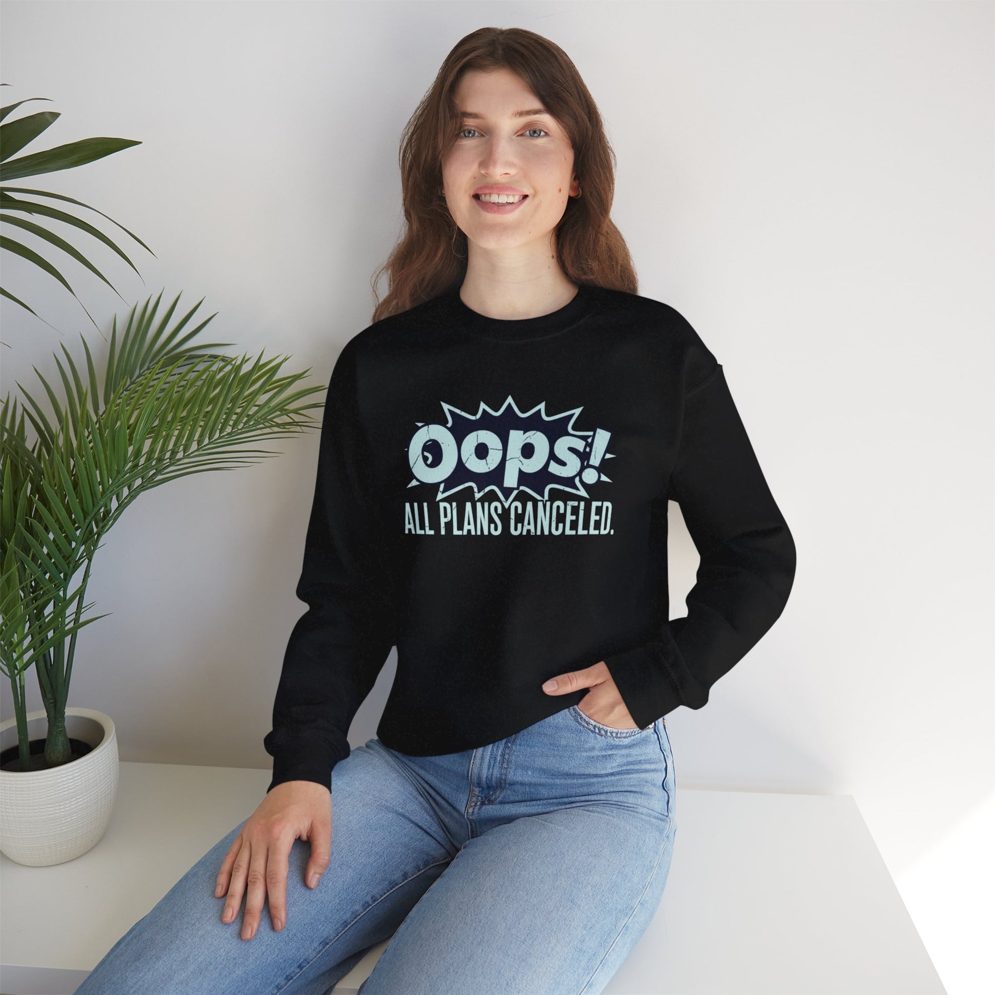 Person wearing a black sweatshirt with 'Oops! All Plans Canceled' text, sitting on a white surface with a plant in the background.