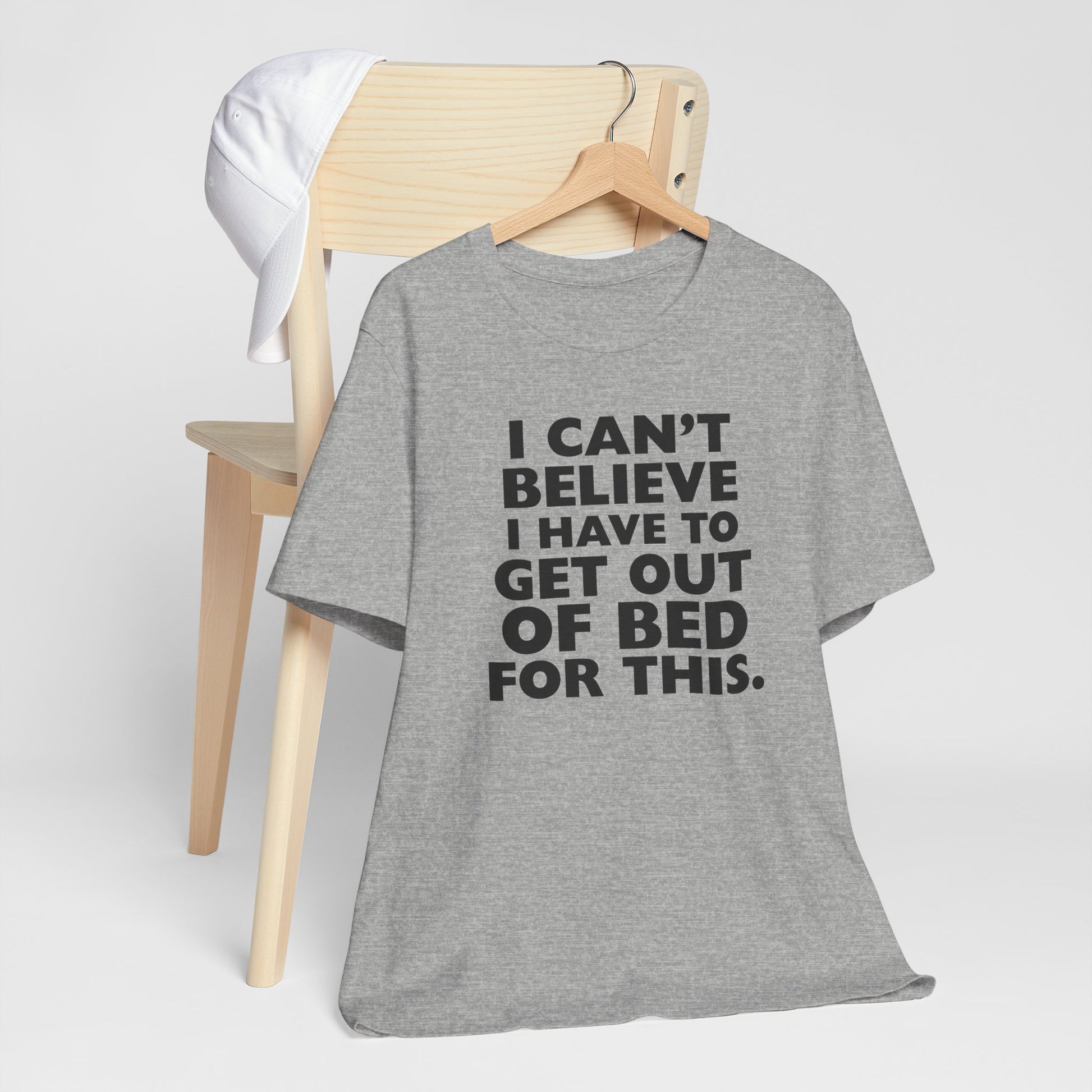 Gray t-shirt with humorous text 'I can't believe I have to get out of bed for this' on a hanger against a white background