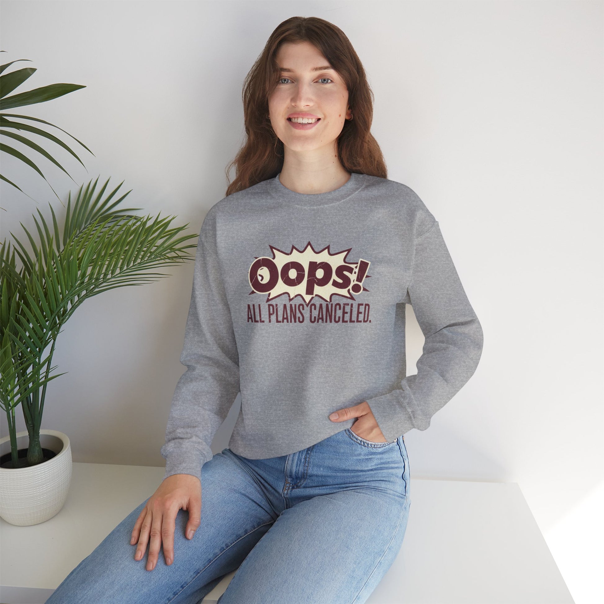 Person wearing a gray sweatshirt with 'Oops! All plans canceled' text, sitting on a white surface with a plant in the background.