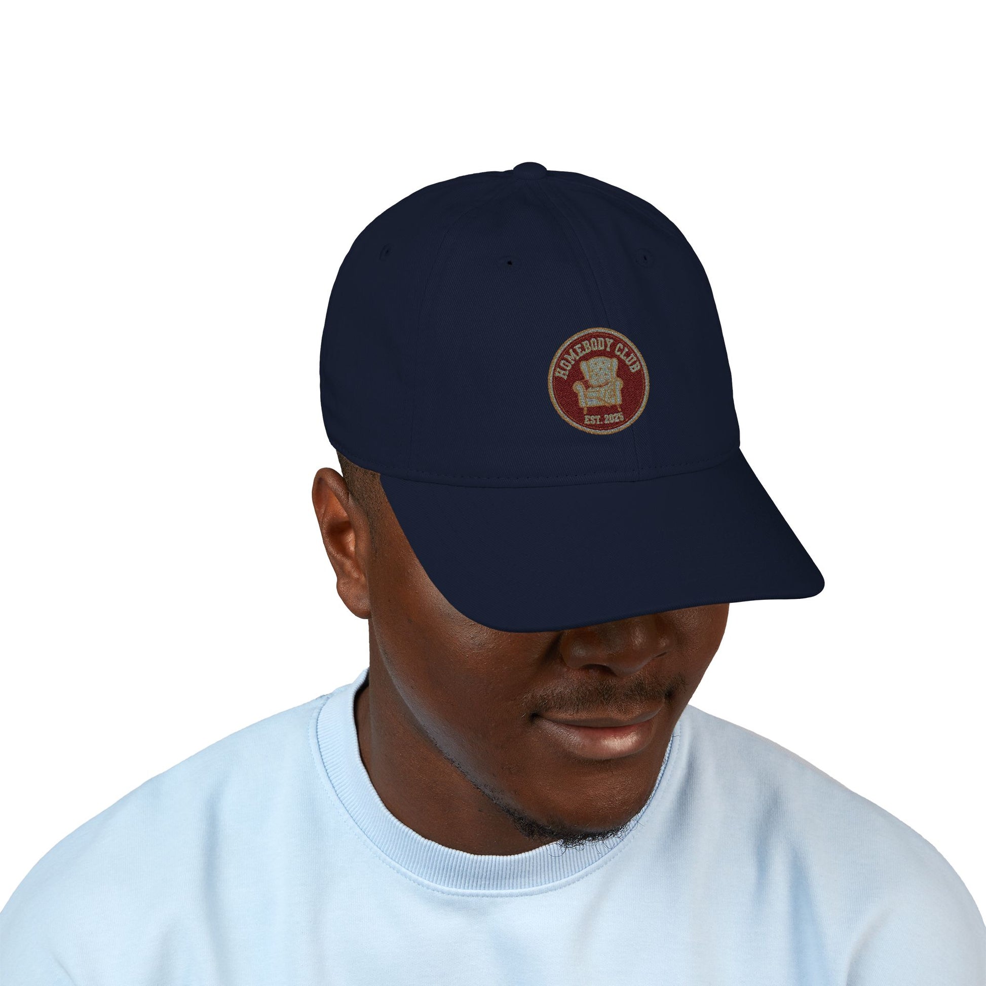 Navy cap with a logo and text 'Homebody club Est. 2025' worn by a person on a white background