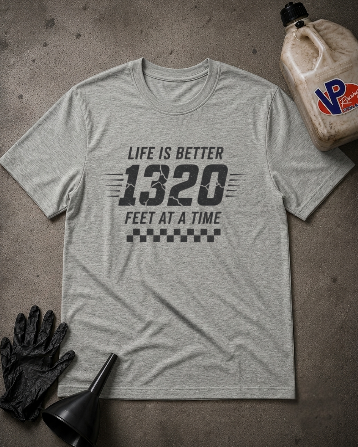 A light heather gray graphic t-shirt laid flat on a textured gray surface, featuring the text 'LIFE IS BETTER 1320 FEET AT A TIME' with a checkered flag motif. The shirt is styled with black mechanic gloves, a black funnel, and a plastic jug of vintage racing fuel in the corners.