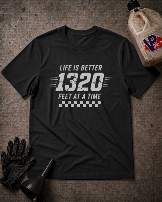 A black graphic t-shirt laid flat on a textured gray surface, featuring the white text 'LIFE IS BETTER 1320 FEET AT A TIME' with a checkered stripe design. The shirt is styled with a black glove, a funnel, and a plastic jug in a garage-themed flat lay.