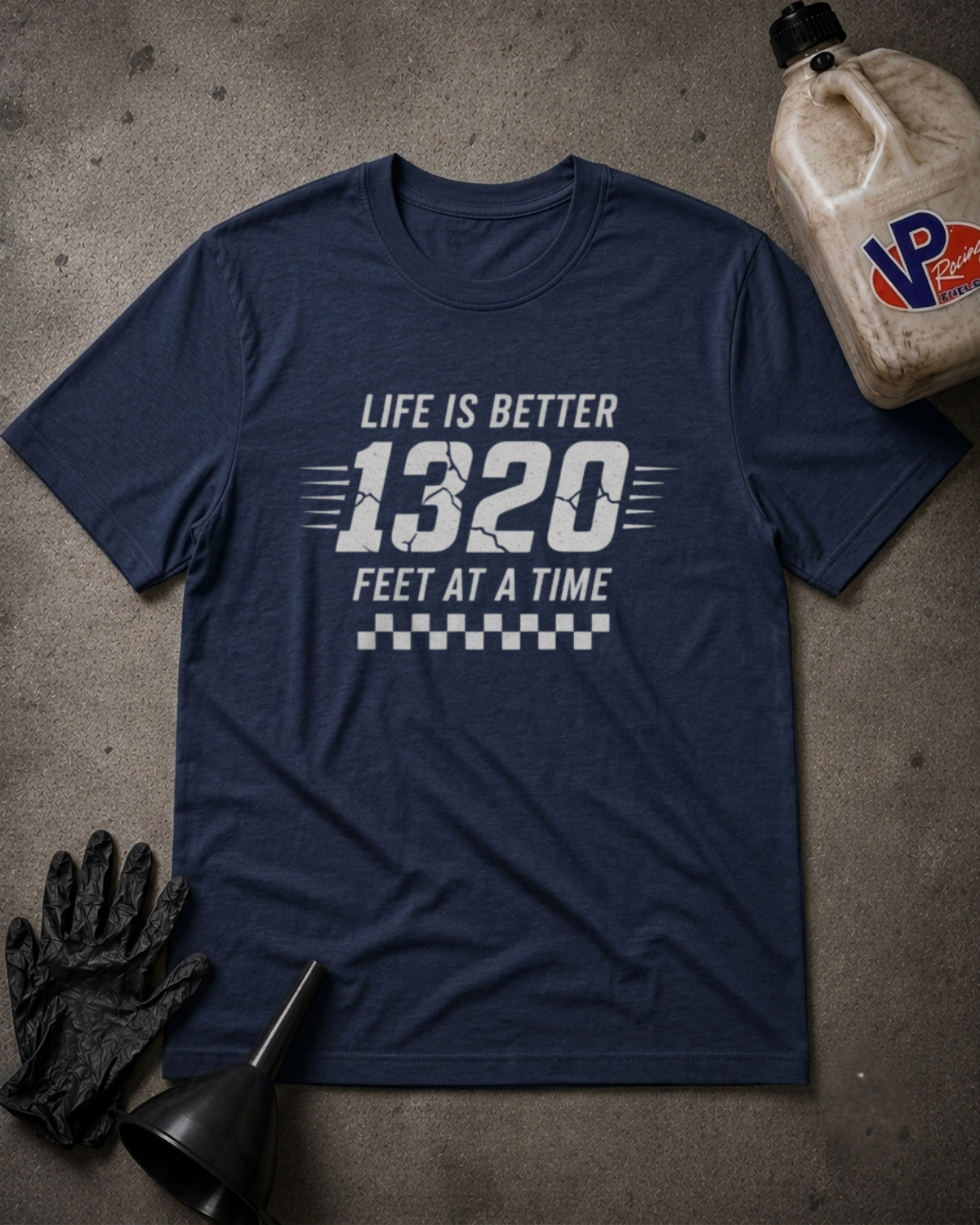A navy blue graphic t-shirt laid flat on a textured gray surface, featuring the white text 'LIFE IS BETTER 1320 FEET AT A TIME' with a checkered stripe design. Black gloves and a funnel are placed near the shirt, and a plastic jug is in the upper right corner.