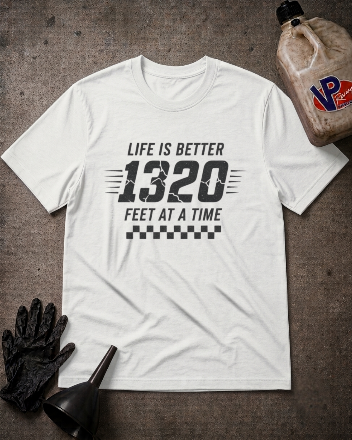 A white graphic t-shirt laid flat on a textured gray surface, featuring the text 'LIFE IS BETTER 1320 FEET AT A TIME' in black. The shirt is styled with a black oil pan, a black funnel, a black glove, and a plastic jug of oil.