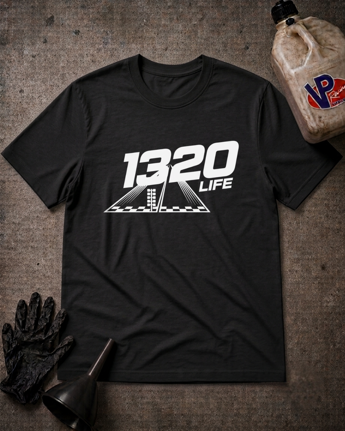 A black t-shirt with a black '1320 LIFE' graphic printed on the chest, styled in a flat-lay with black mechanic gloves, a funnel, and a plastic jug on a textured gray concrete floor.