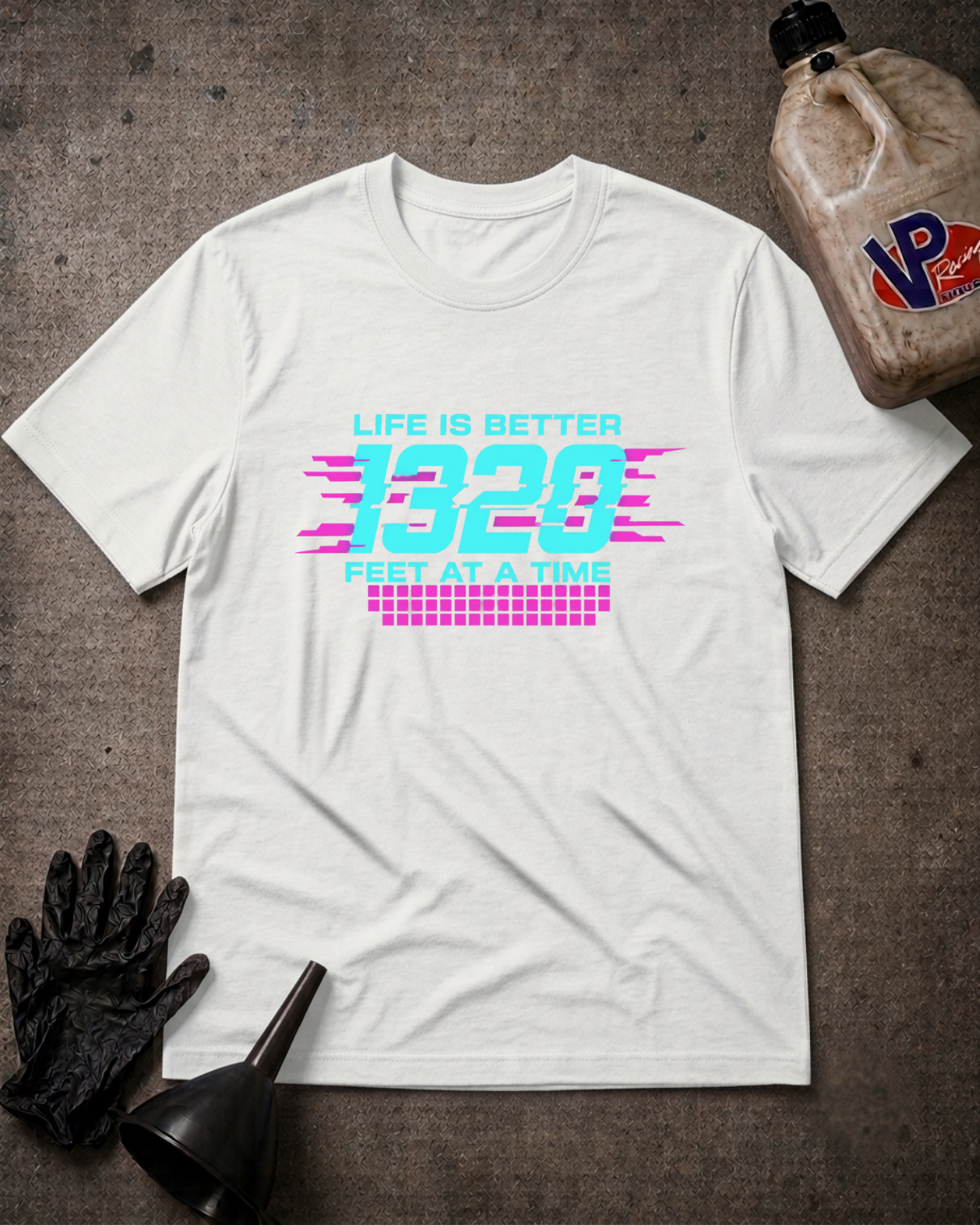 A white short-sleeve graphic T-shirt laid flat on a dark textured surface, surrounded by black gloves, a black funnel, and a brown plastic jug. The shirt has a colorful retro-style chest print reading “LIFE IS BETTER 1320 FEET AT A TIME” in cyan and pink.